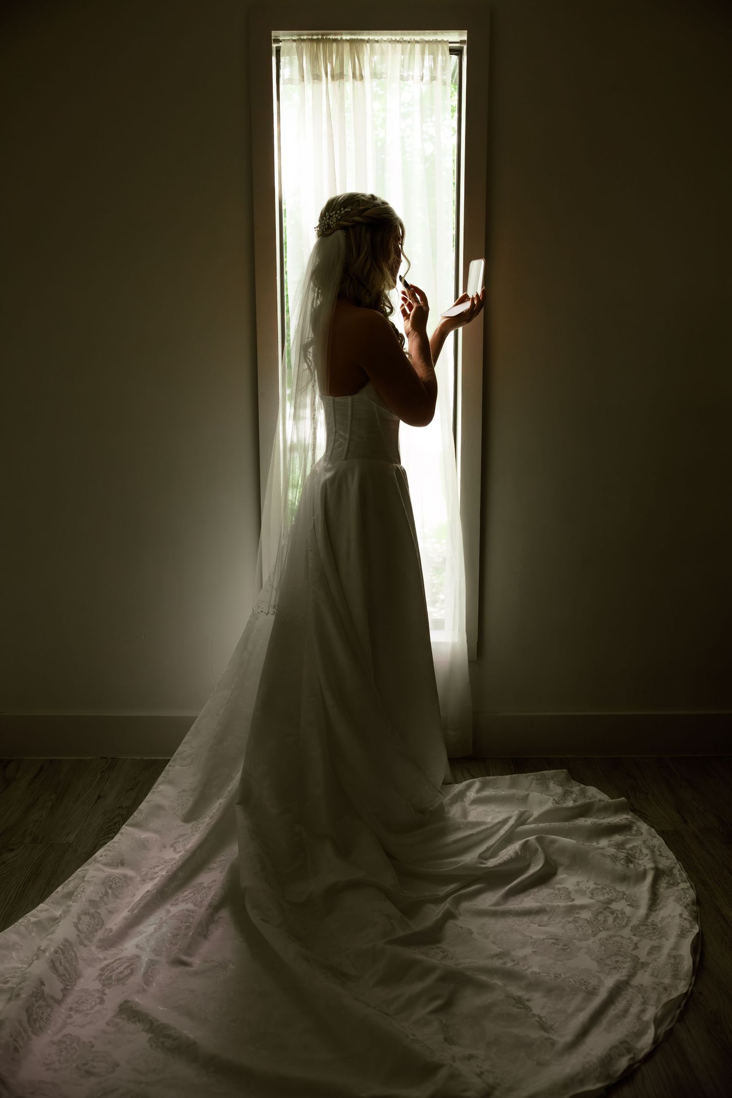 A bride in a flowing white wedding dress stands silhouetted against a bright window in a dark room.