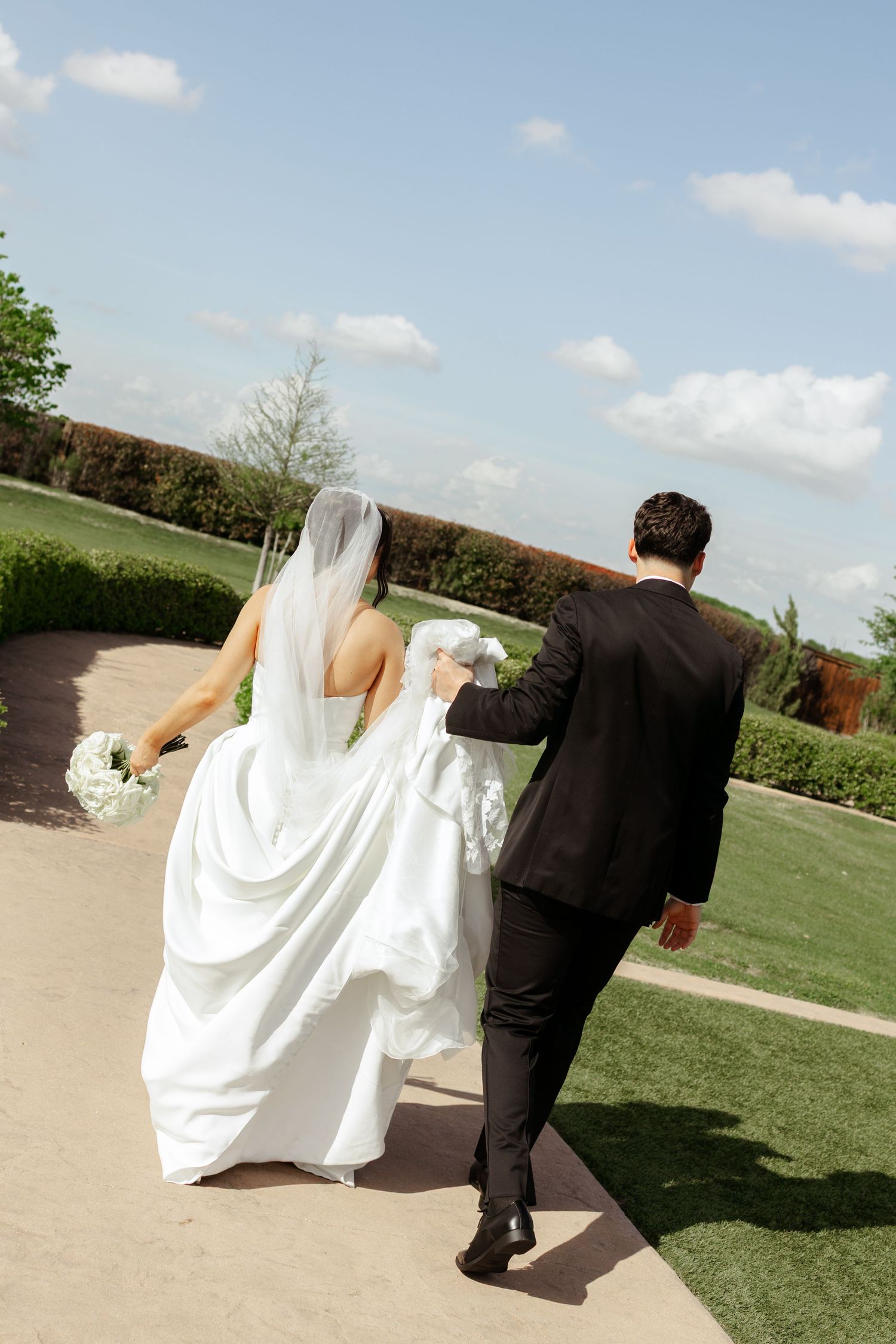 Bride and groom walking hand in hand outdoors, bride in white gown with veil, groom in black suit.