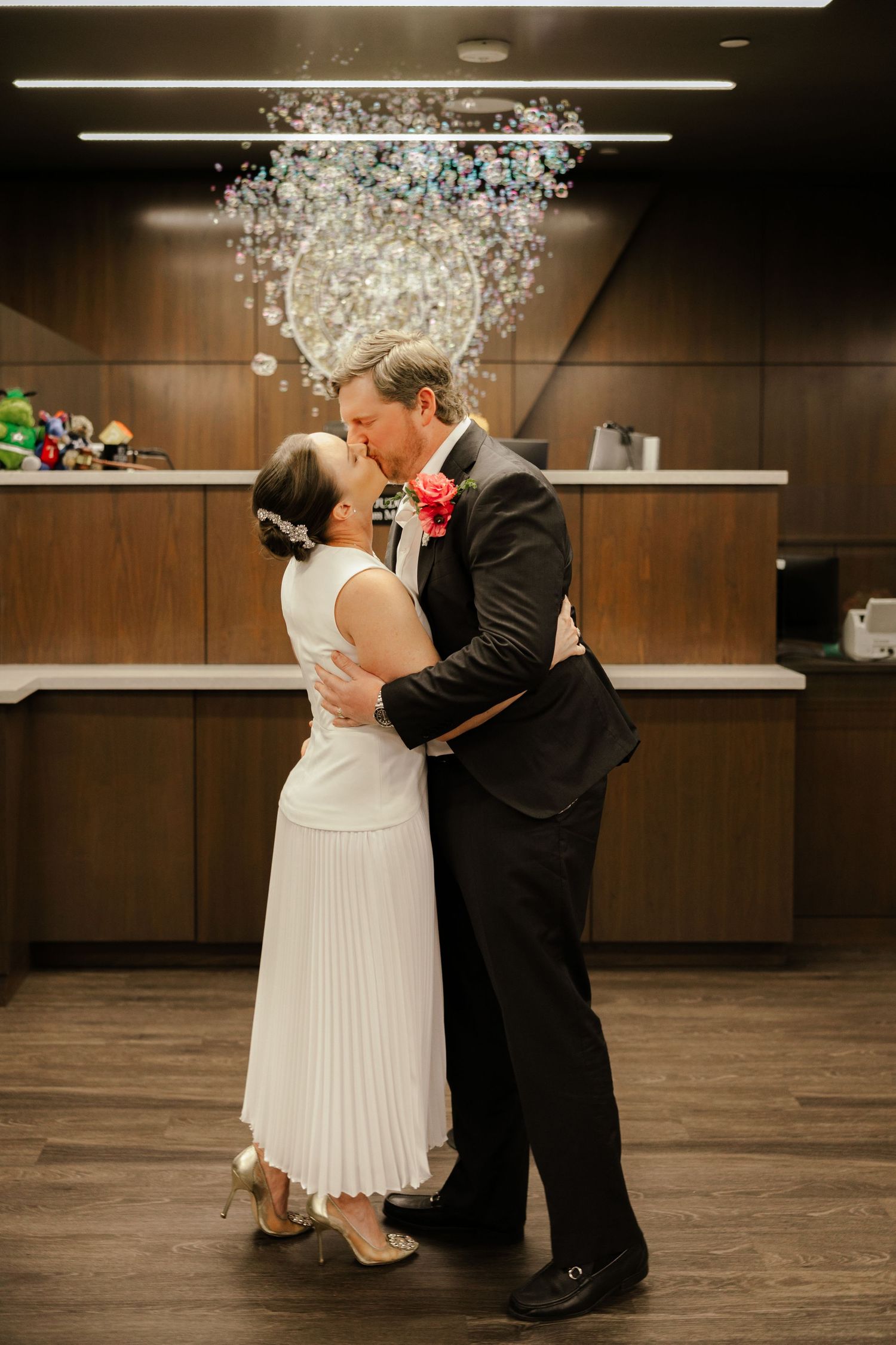 Bride and groom share first kiss in modern venue with crystal chandelier, bride in white pleated dress, groom in black suit.