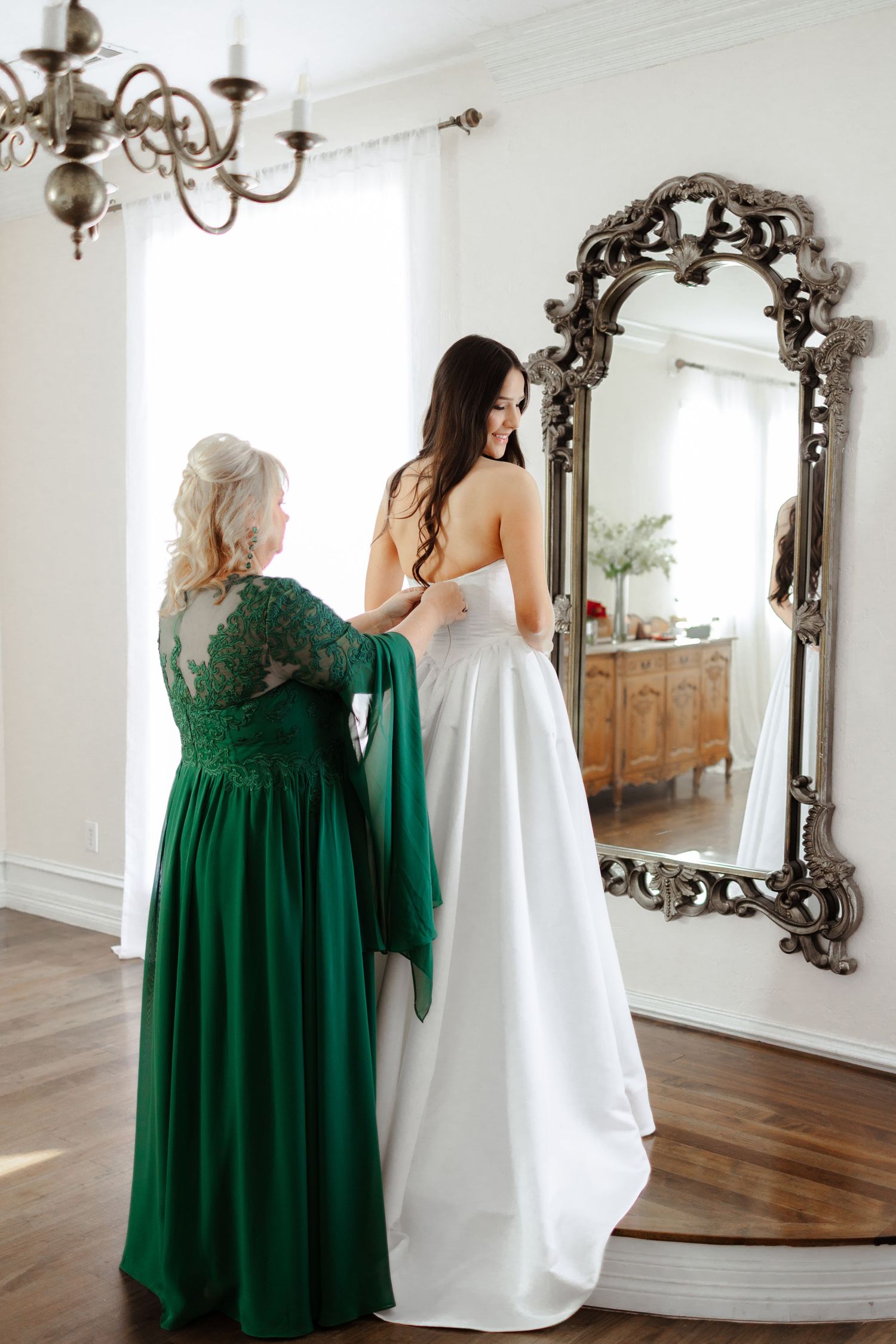 A person in an emerald green dress helps adjust a white wedding gown in a room with ornate mirrors and wooden floors.