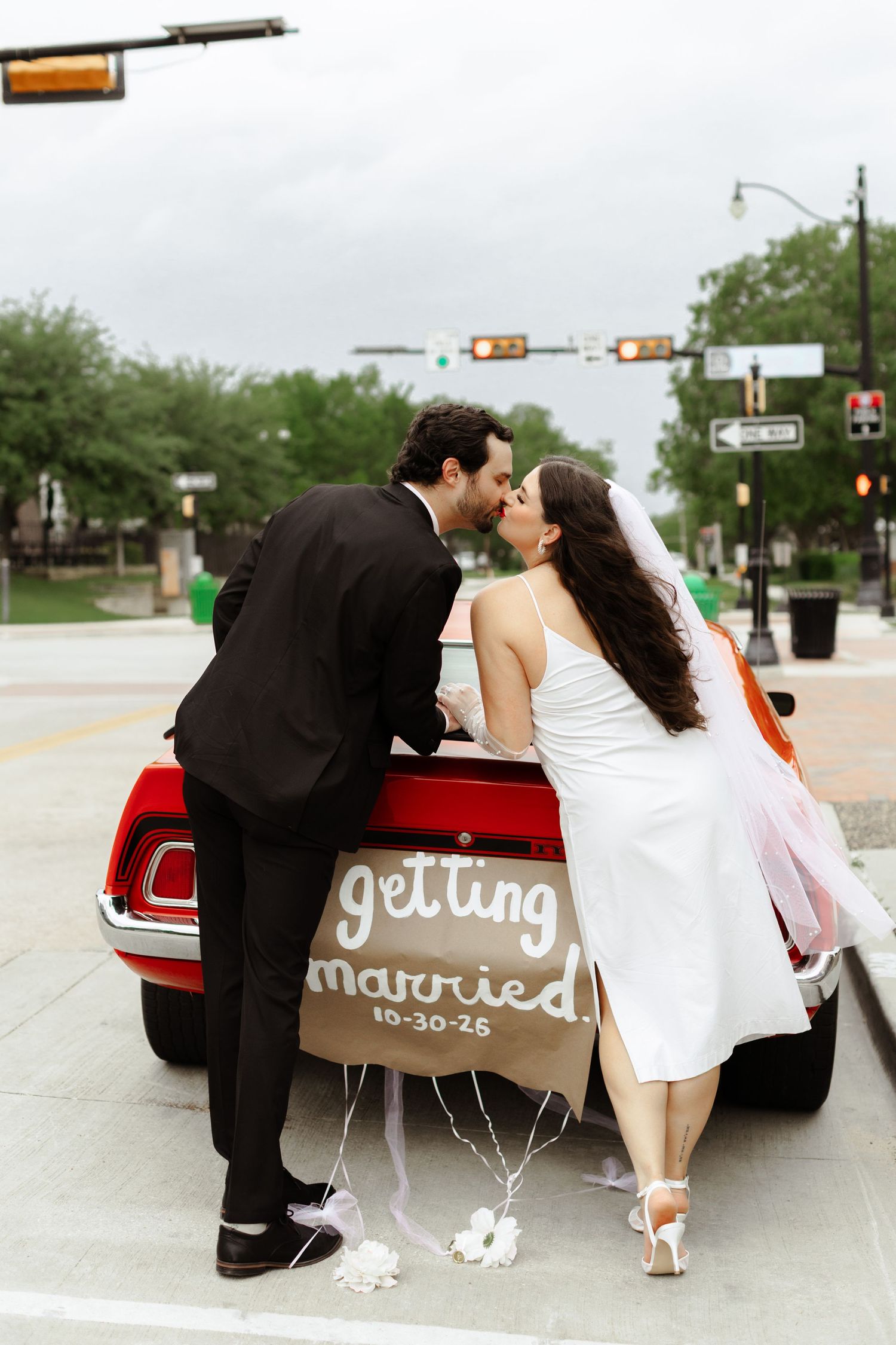 Newlyweds kissing beside a red vintage car with a getting married sign dated 10-30-25 on their wedding day.