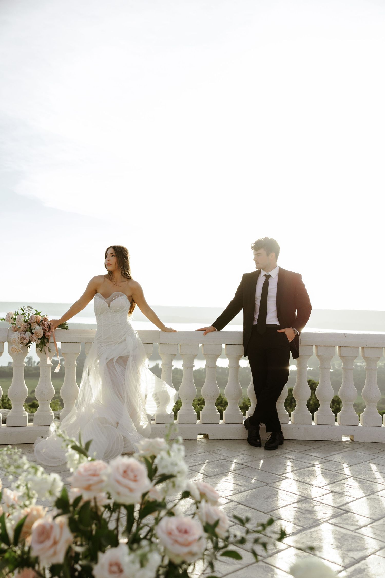 Bride and groom hold hands on a white balustrade terrace overlooking the sea, surrounded by elegant floral arrangements.