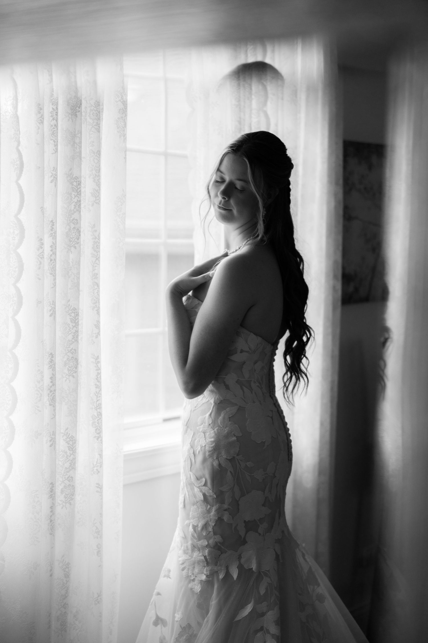 Elegant bride in lace mermaid wedding dress standing by window in black and white bridal portrait.