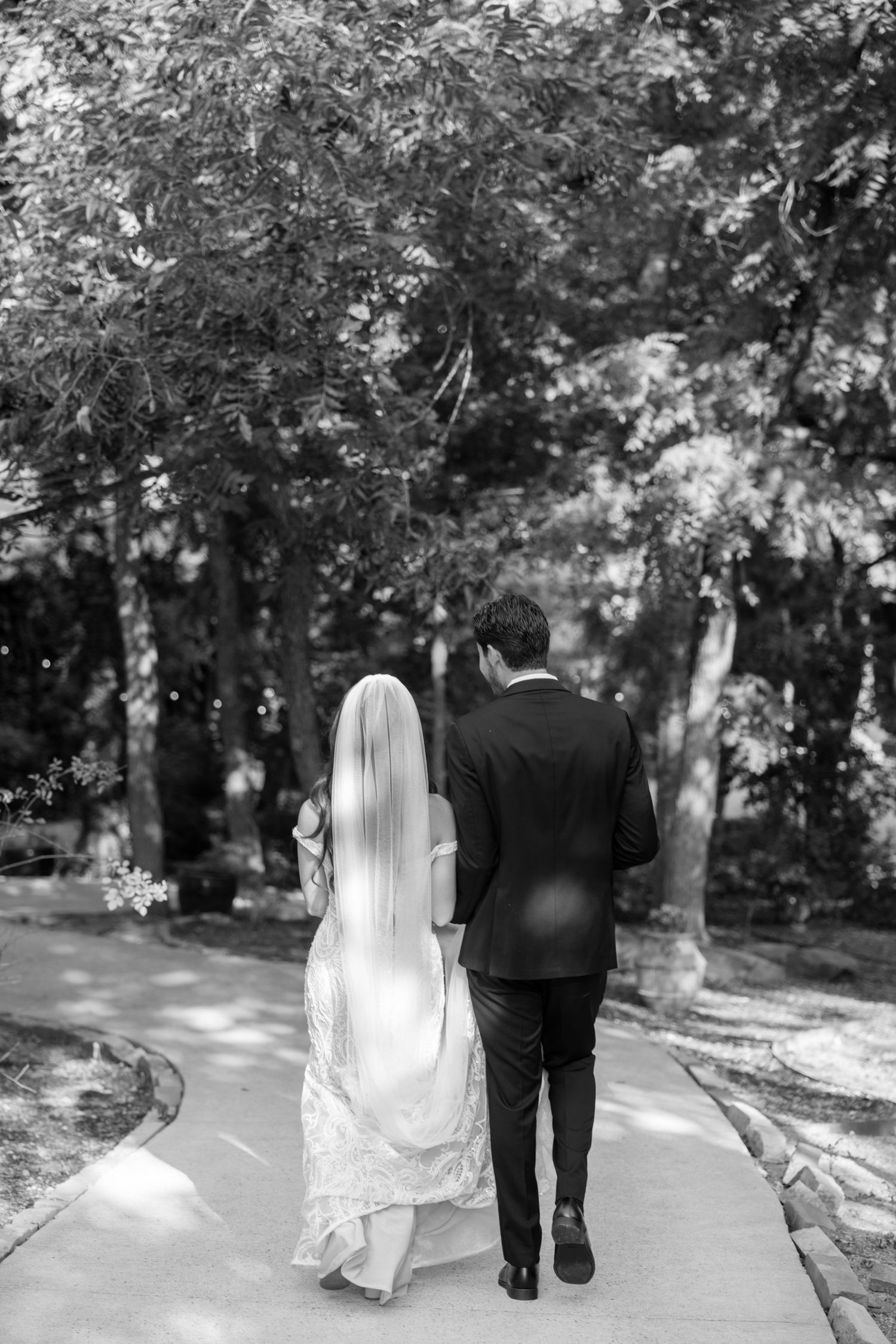 Black and white photo of bride and groom walking hand in hand down a tree-lined path on their wedding day.