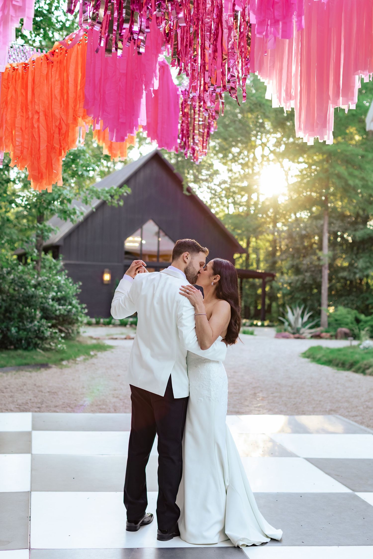 A romantic outdoor wedding moment under vibrant pink and orange streamers at sunset.