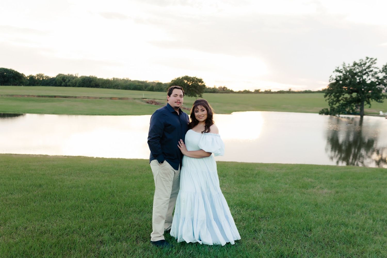 Couple embracing near peaceful pond with green fields in background.