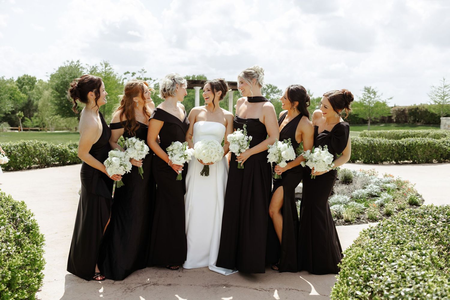 Bride in white gown with bridesmaids in black dresses holding white bouquets in an outdoor garden setting.