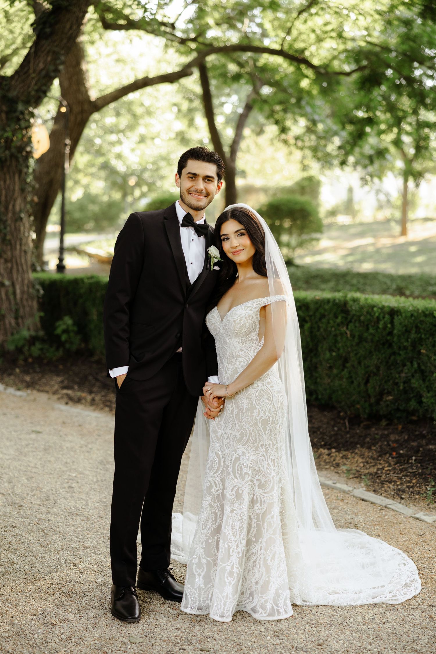 Bride in lace gown and veil stands with groom in black tuxedo outdoors among lush green trees on wedding day.