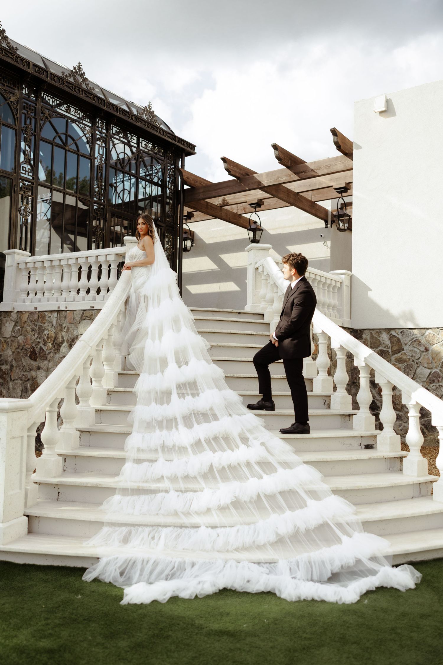 Bride in stunning ruffled white gown with long train poses with groom on elegant white marble staircase.