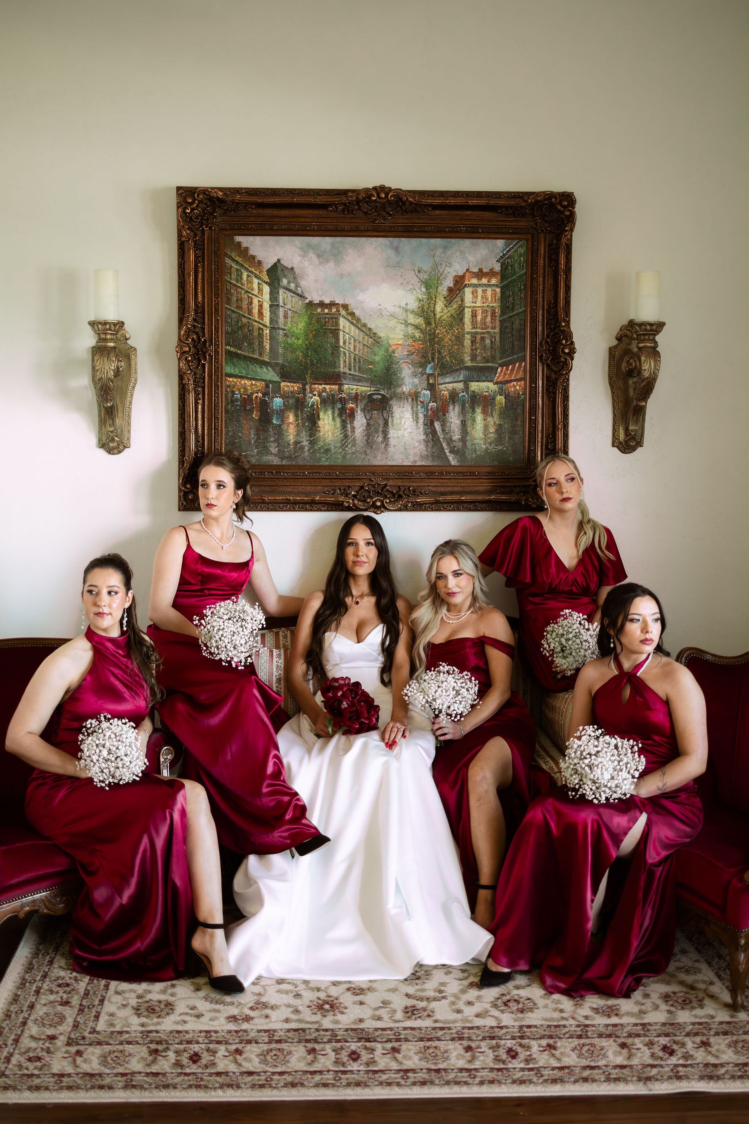 A wedding party in burgundy dresses and white gown sit together on an ornate couch beneath framed artwork.