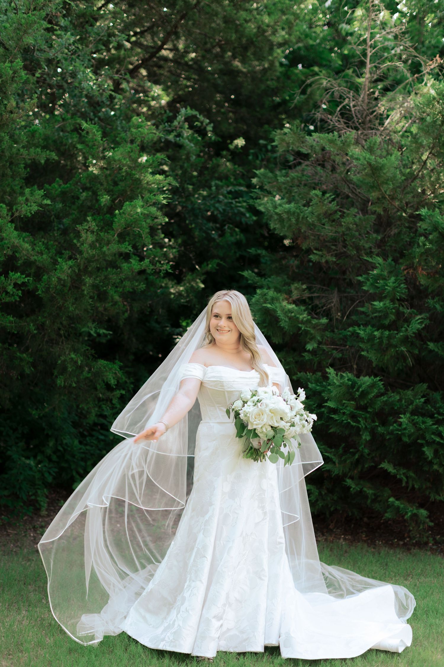 Bride in flowing white dress and veil poses gracefully on grass with greenery backdrop.