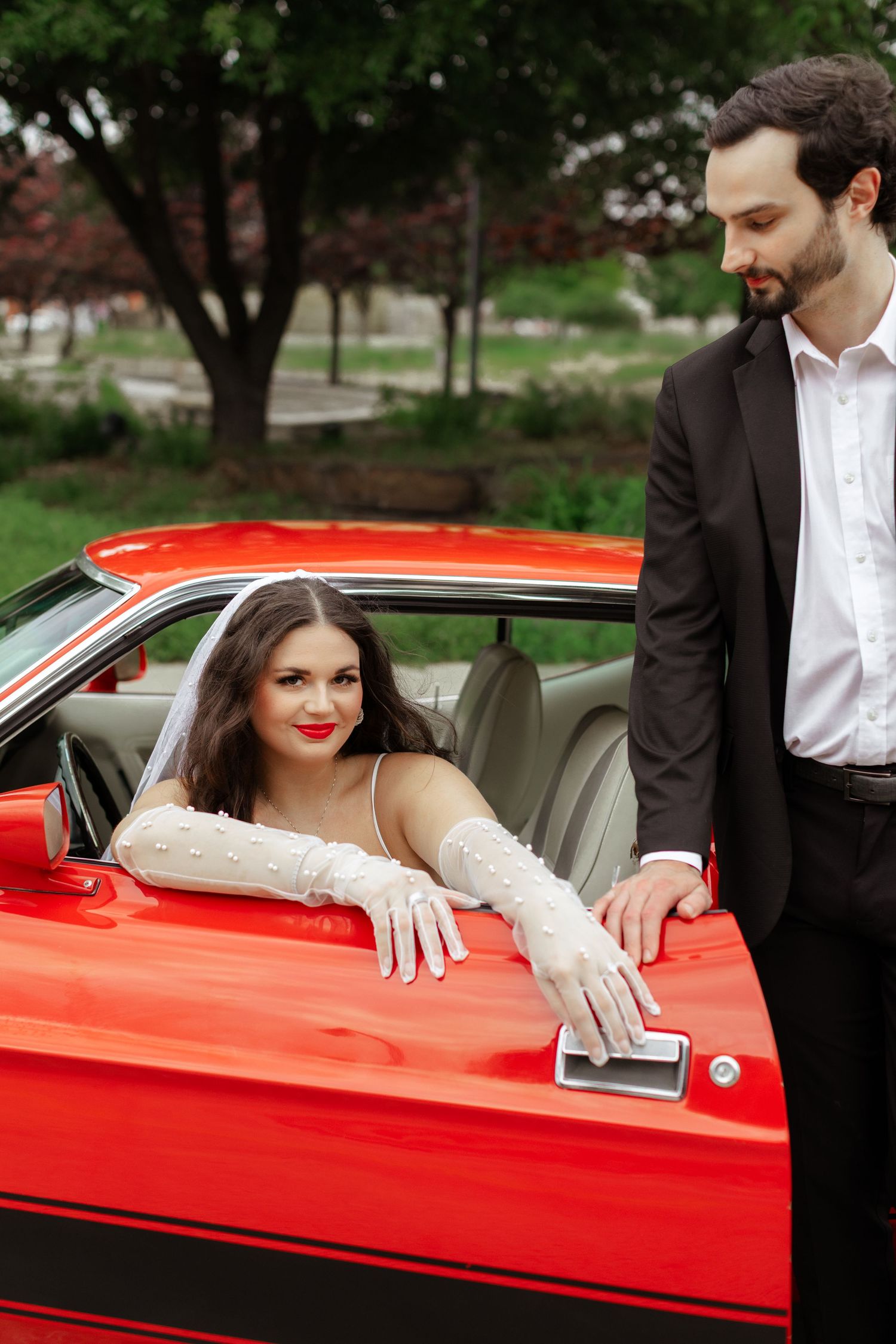 Bride in white dress and gloves sits in red classic convertible car as groom in black suit opens door.