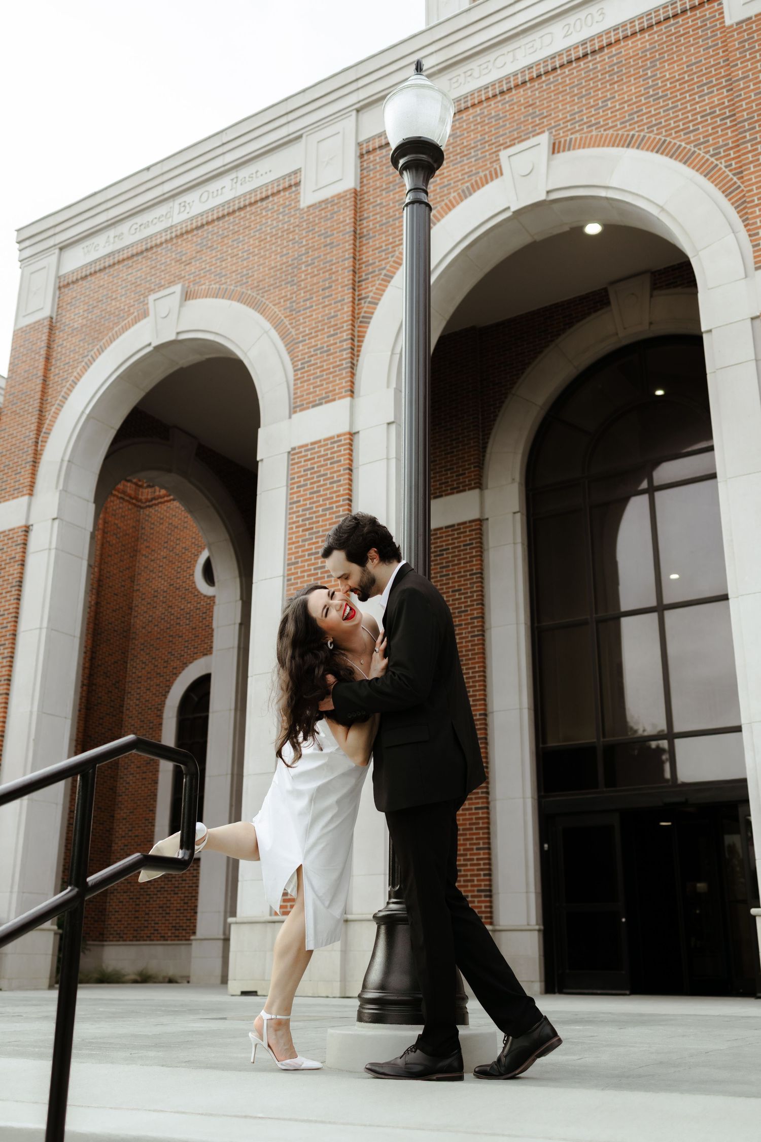 Couple sharing a romantic kiss by a lamppost outside a brick building with arched windows.