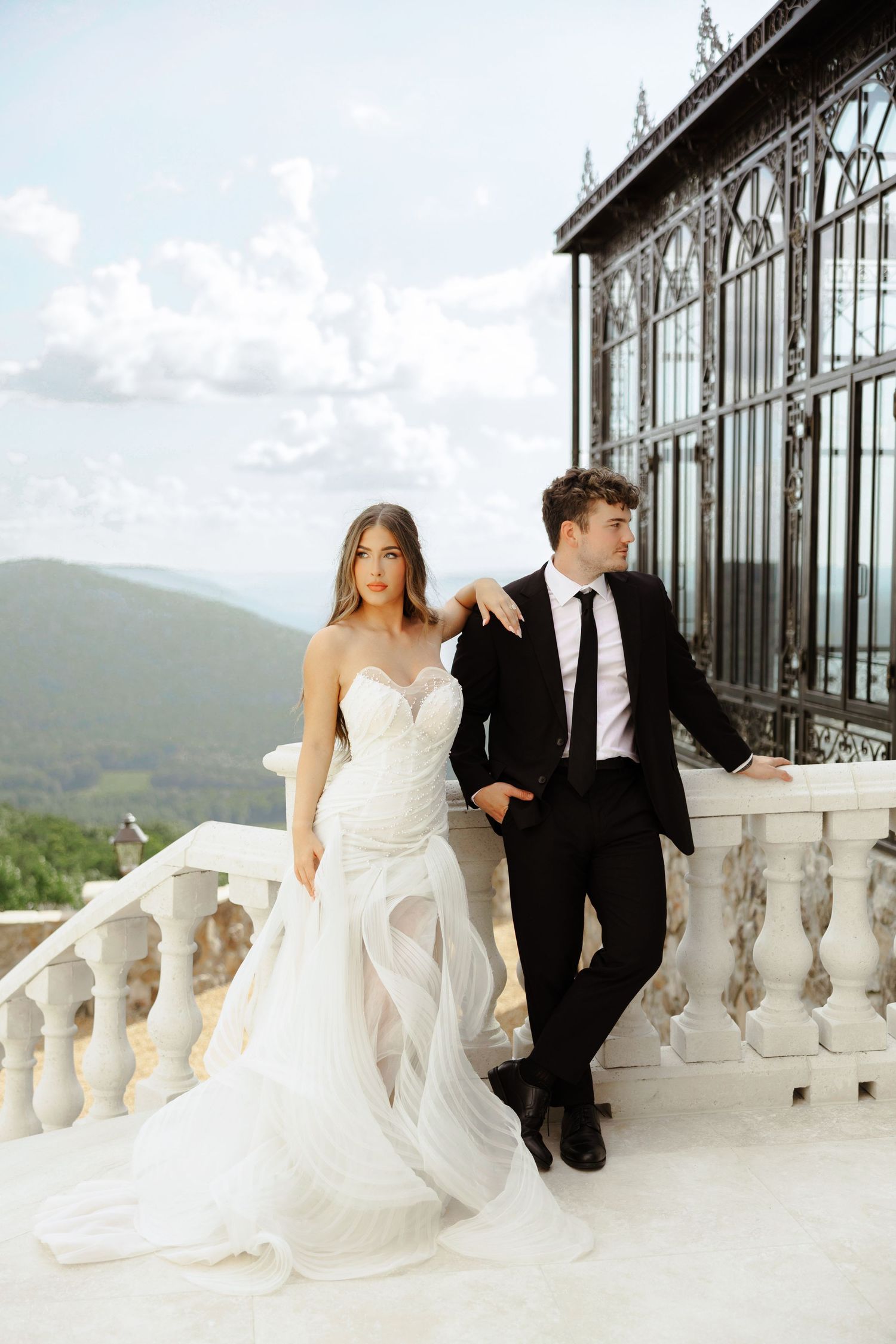 Elegant bride in white strapless gown stands with groom in black suit on a marble balcony with mountain views.