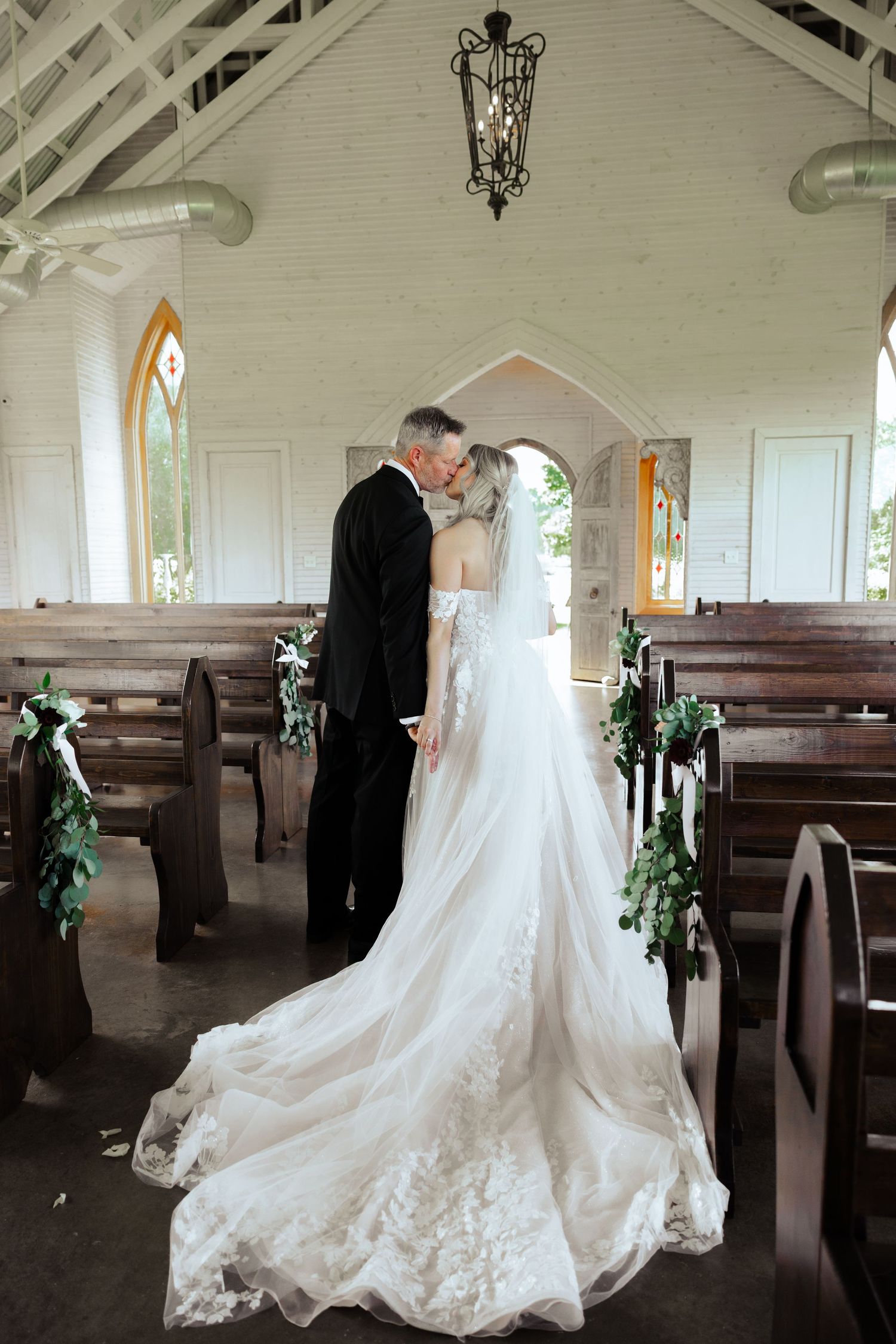 A romantic chapel wedding scene with wooden pews decorated with greenery garlands.
