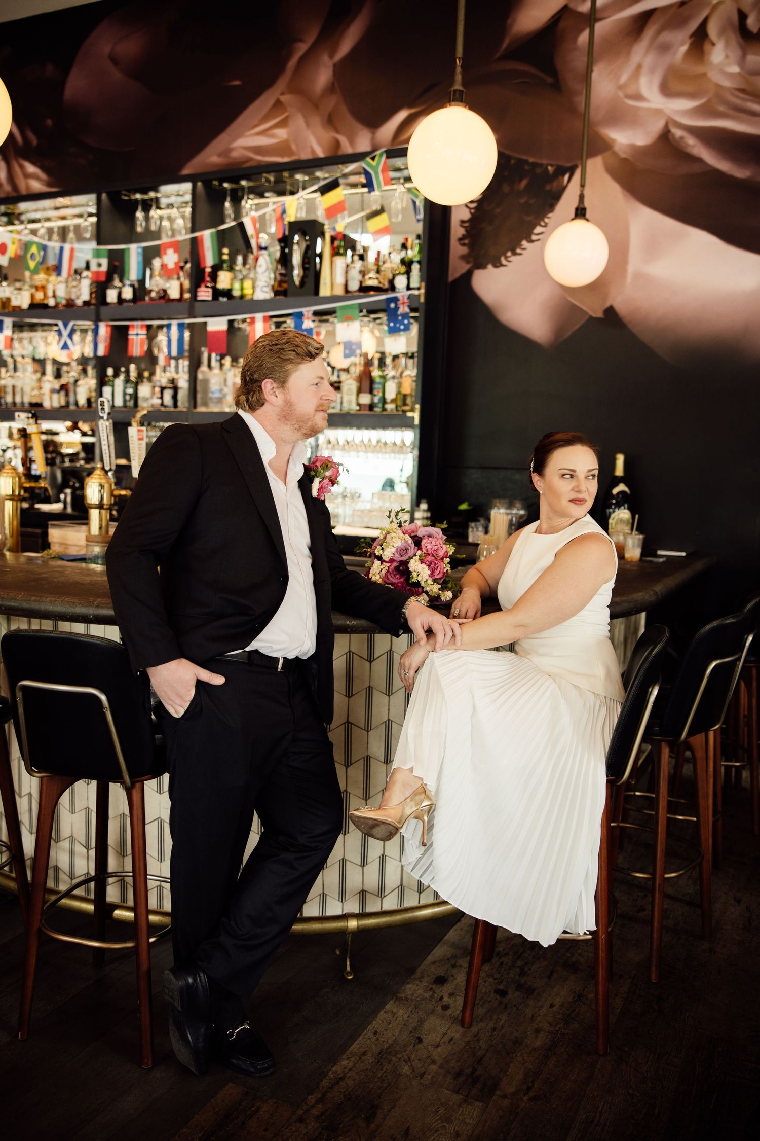 Bride and groom share a romantic moment at a bar, bride in white pleated dress holding pink bouquet.