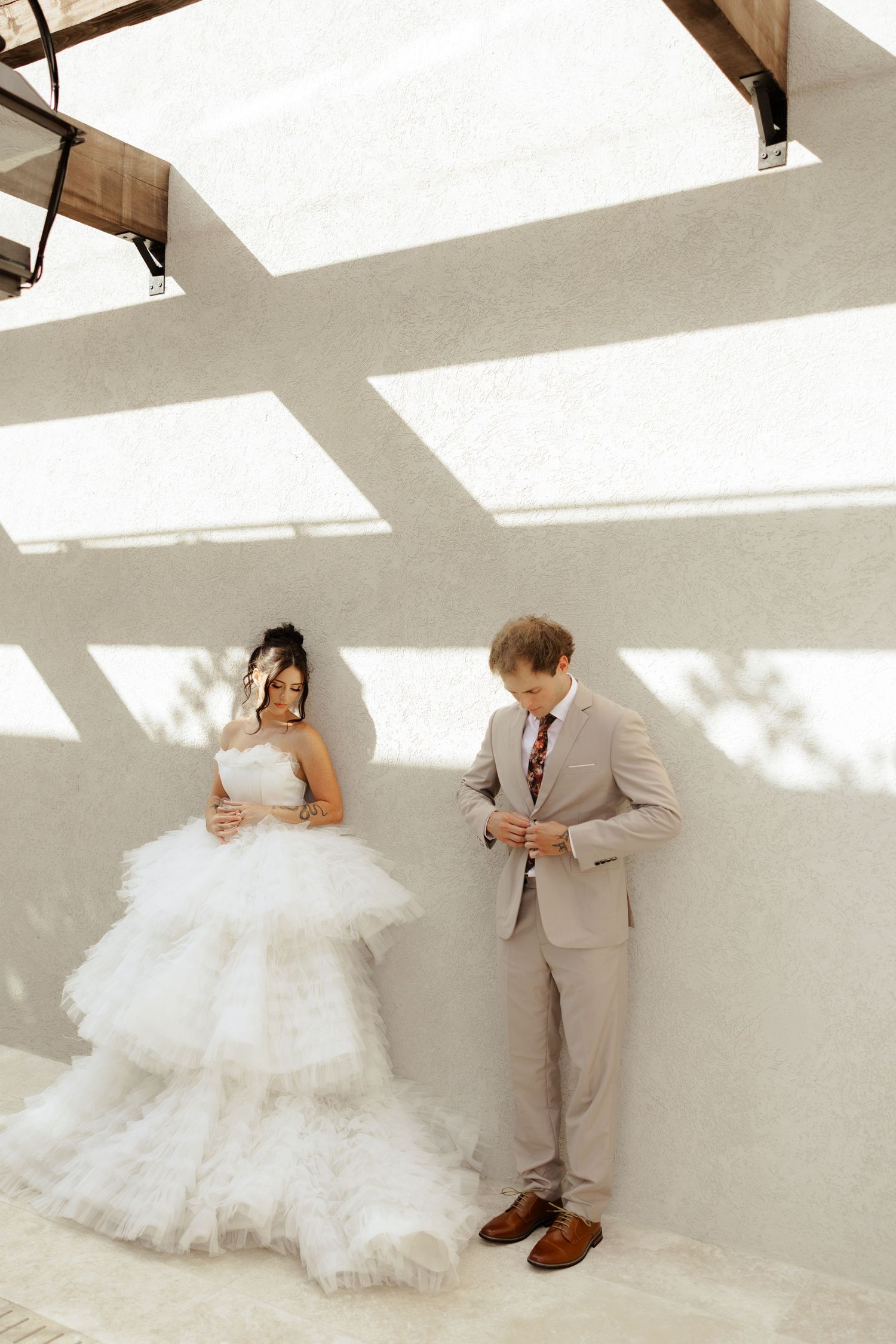 Bride in layered tulle gown and groom in beige suit stand against white wall in elegant wedding portrait.