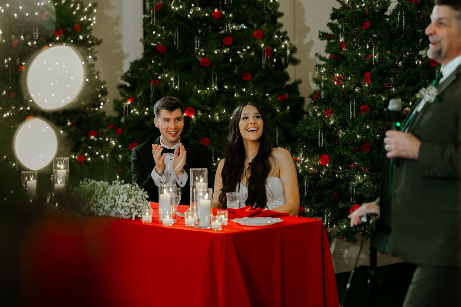 Couple sits at red-clothed table with champagne glasses in front of decorated Christmas trees during holiday celebration.