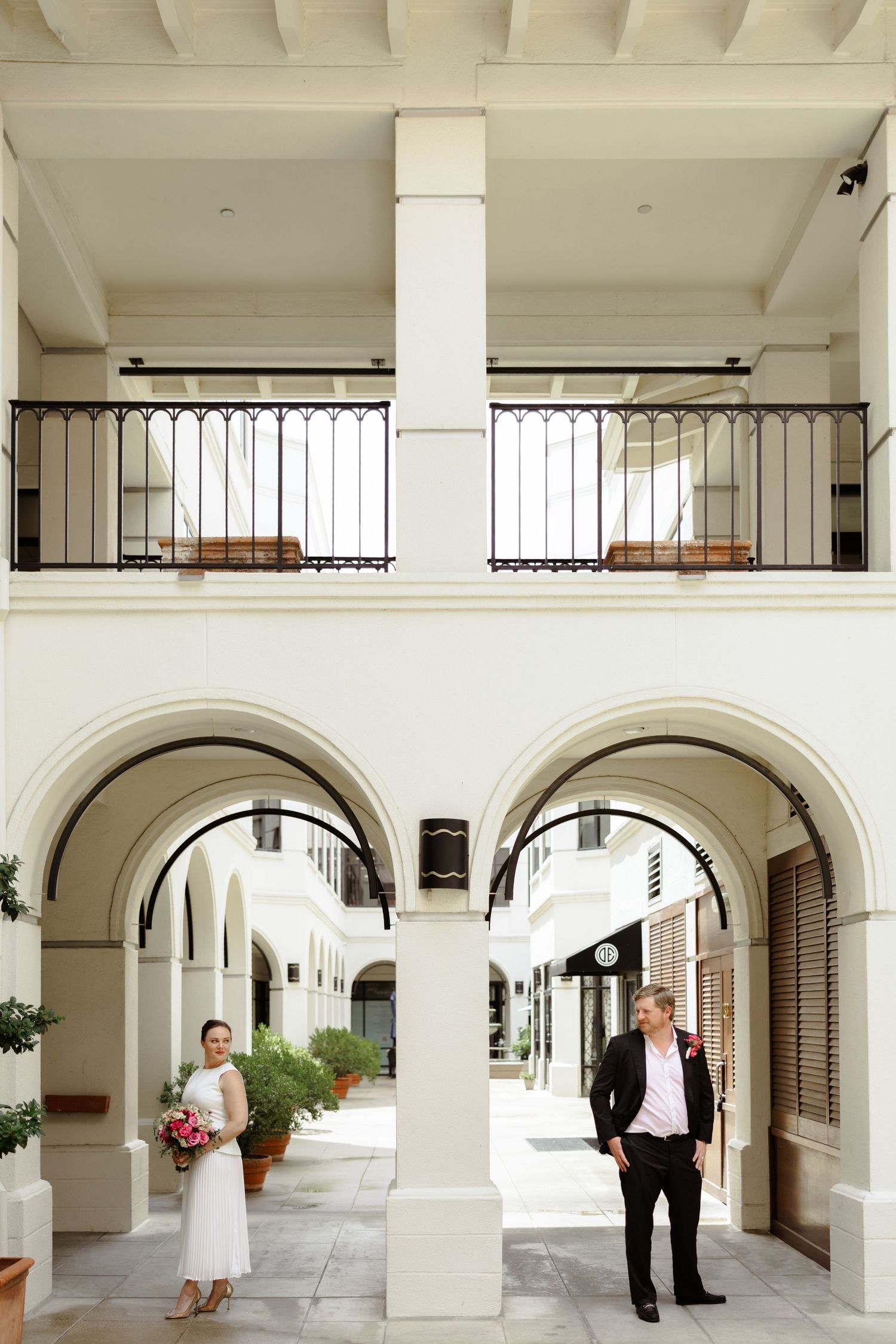 Bride and groom pose under white arched colonnade of elegant two-story building with wrought iron balcony.