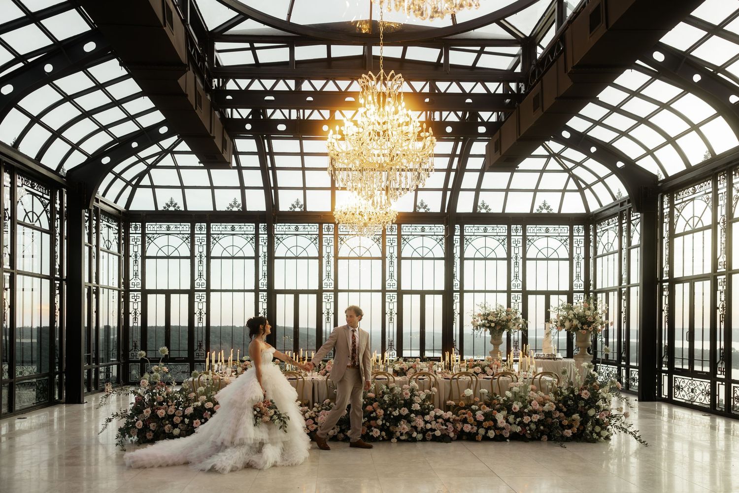 Bride and groom stand beneath a grand chandelier in an elegant glass conservatory wedding venue adorned with lush floral arrangements.