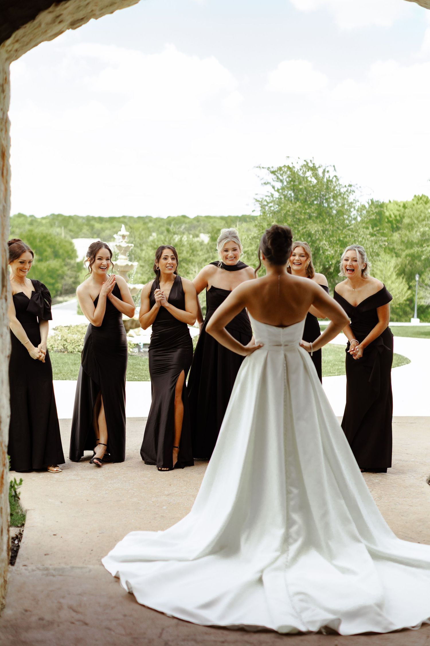 Bride in white gown with train reveals dress to bridesmaids in black dresses outdoors at elegant venue.