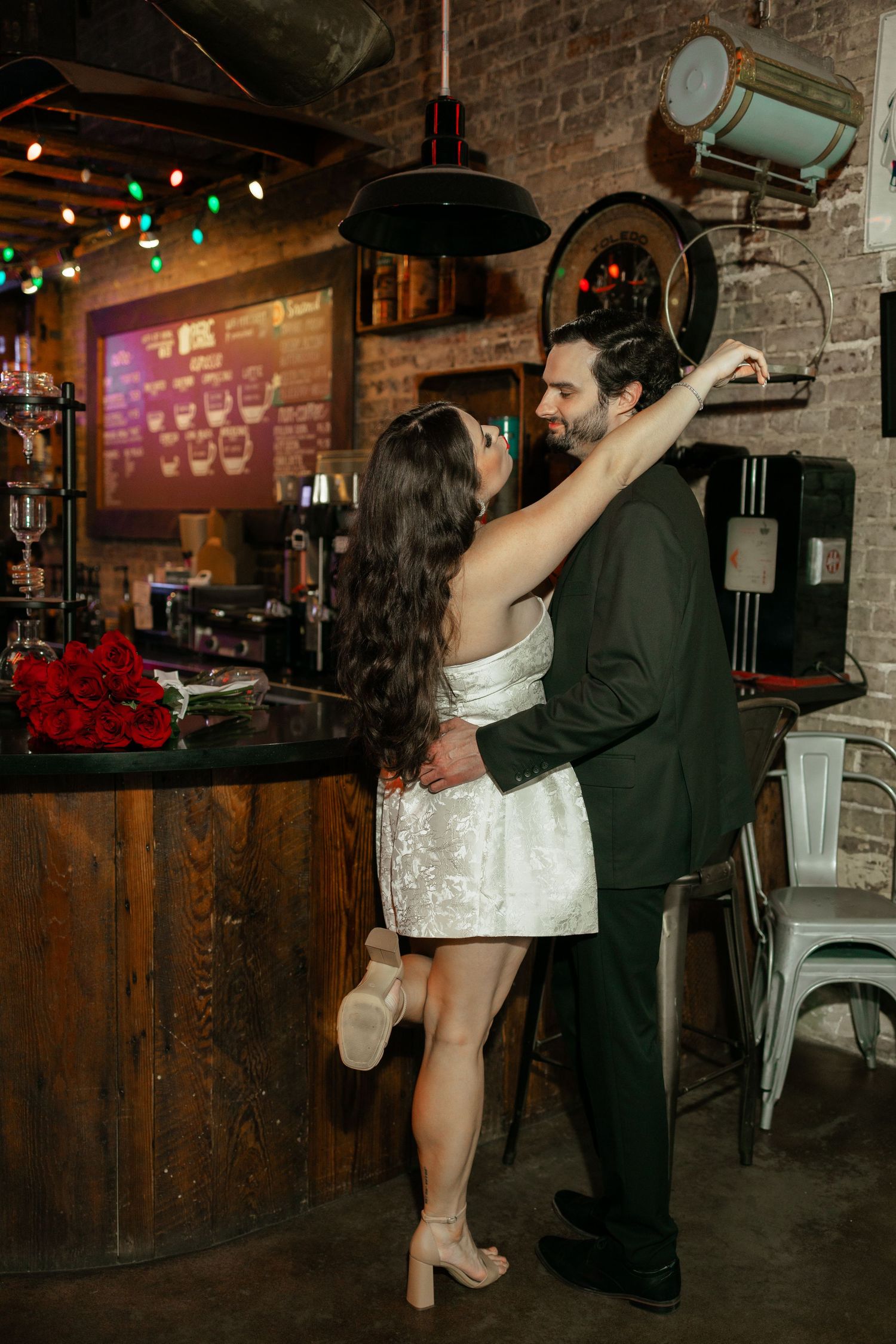 Couple in wedding attire dance romantically at a rustic bar with red roses and warm string lights.