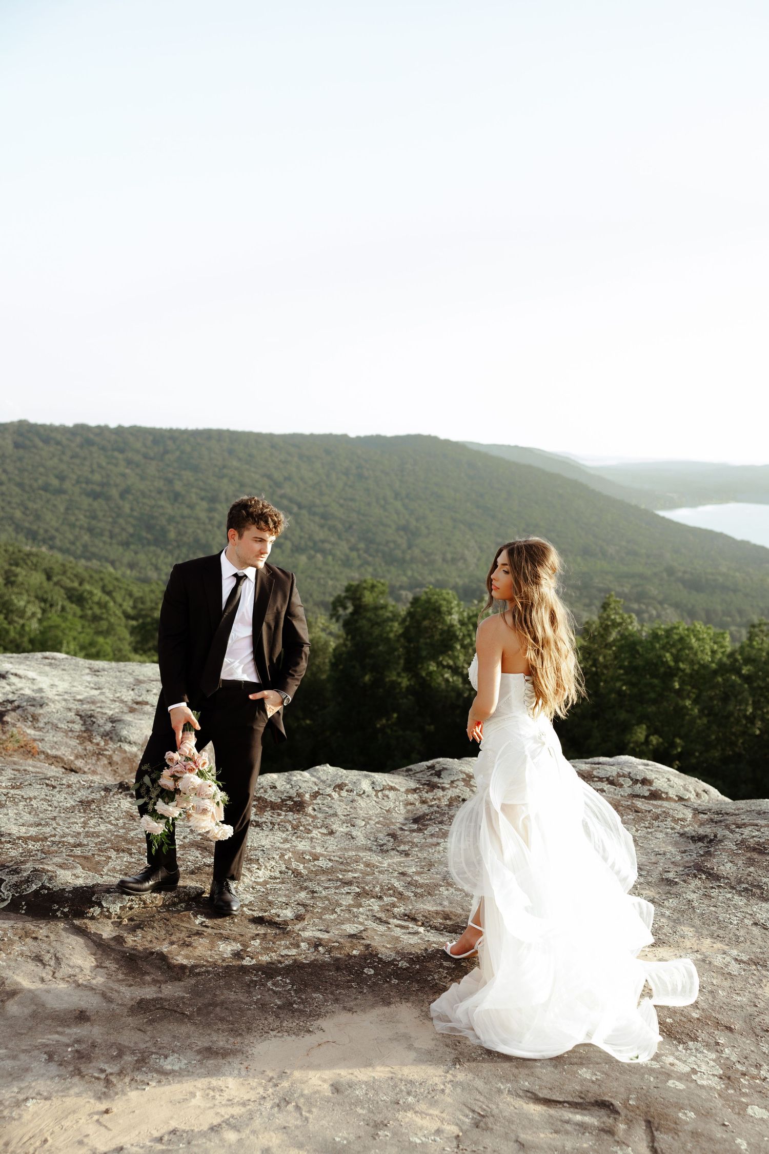 Bride and groom stand on rocky mountain overlook, she in flowing white gown, he in dark suit holding bouquet.
