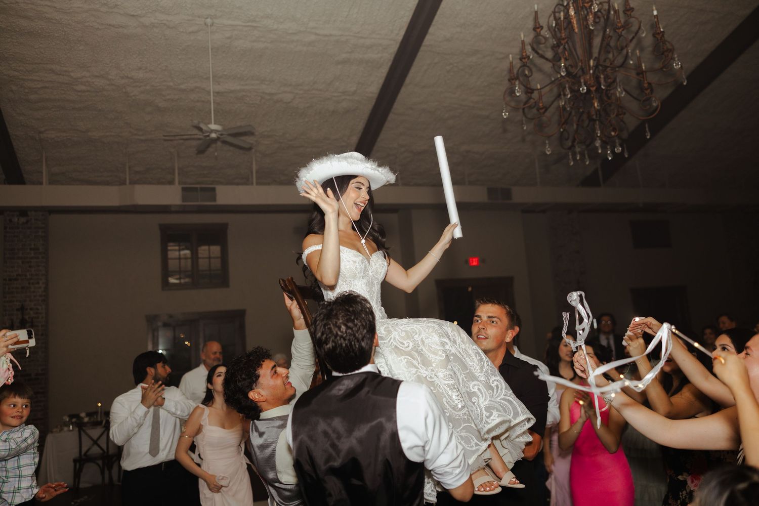 Bride lifted on shoulders during lively wedding reception dance floor celebration with guests cheering around her.