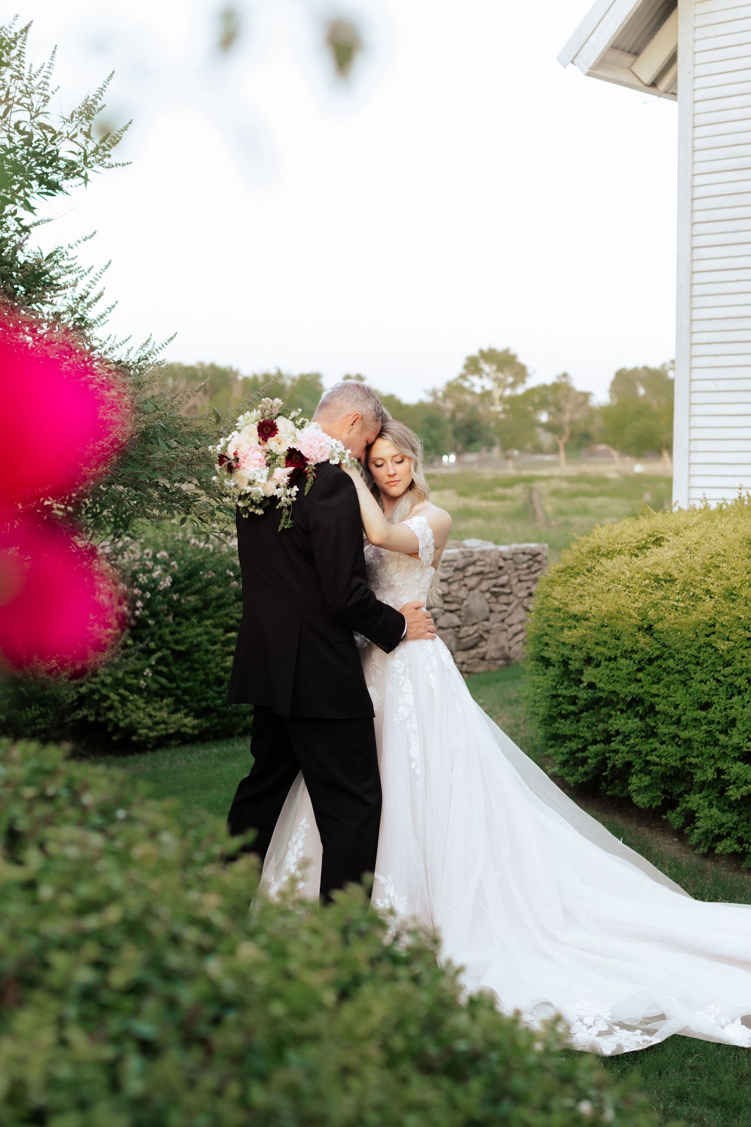A romantic wedding photo featuring a flowing white dress and black suit against lush garden greenery.
