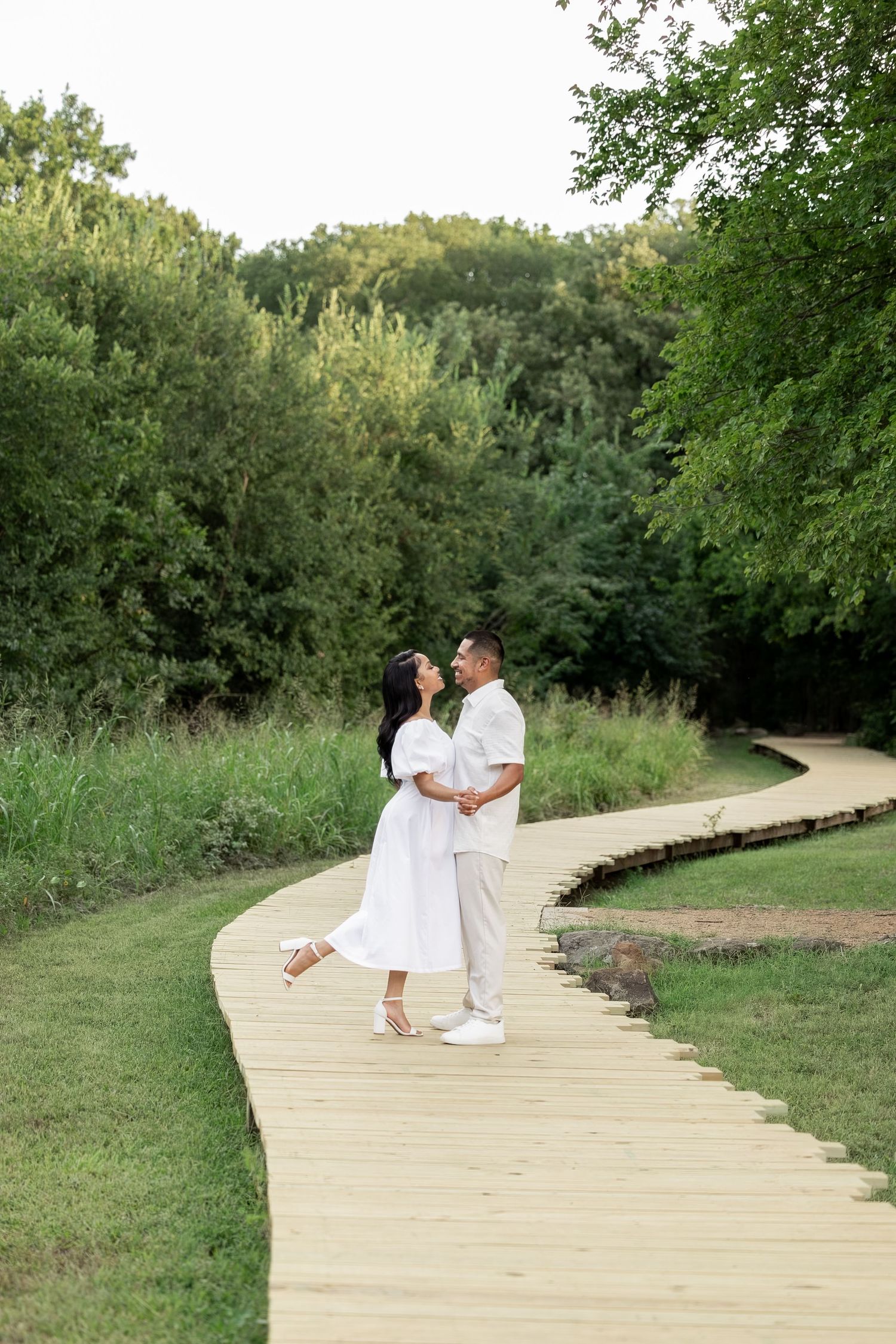 A couple in white outfits shares a romantic moment on a winding wooden boardwalk surrounded by lush greenery.
