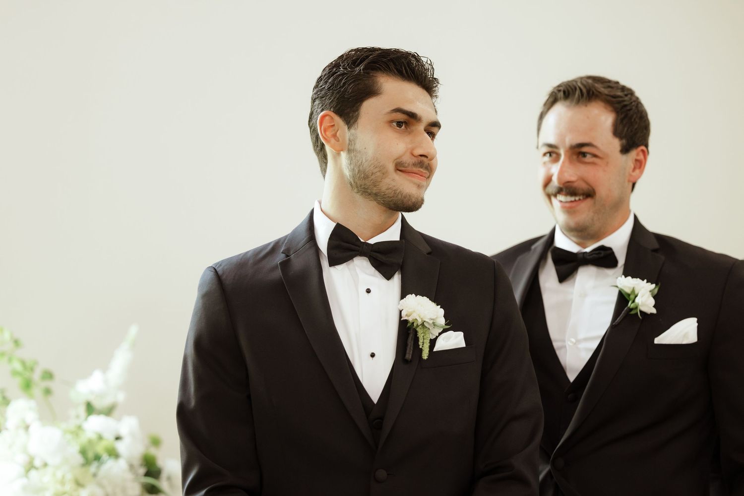 Two men in black tuxedos with bow ties and boutonnieres smile at a formal wedding ceremony.