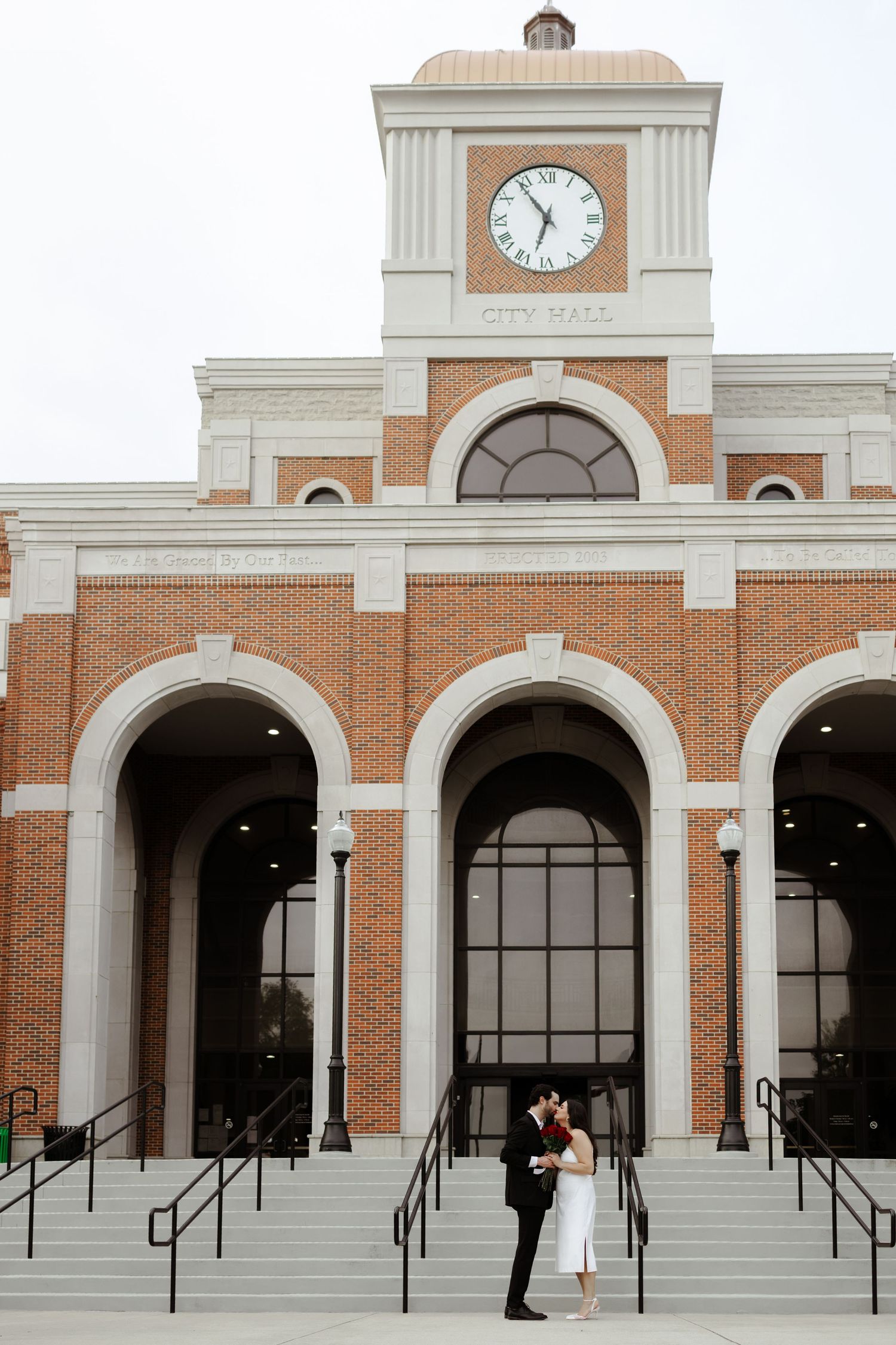 Couple sharing a kiss on the steps of a brick City Hall building with a clock tower on their wedding day.