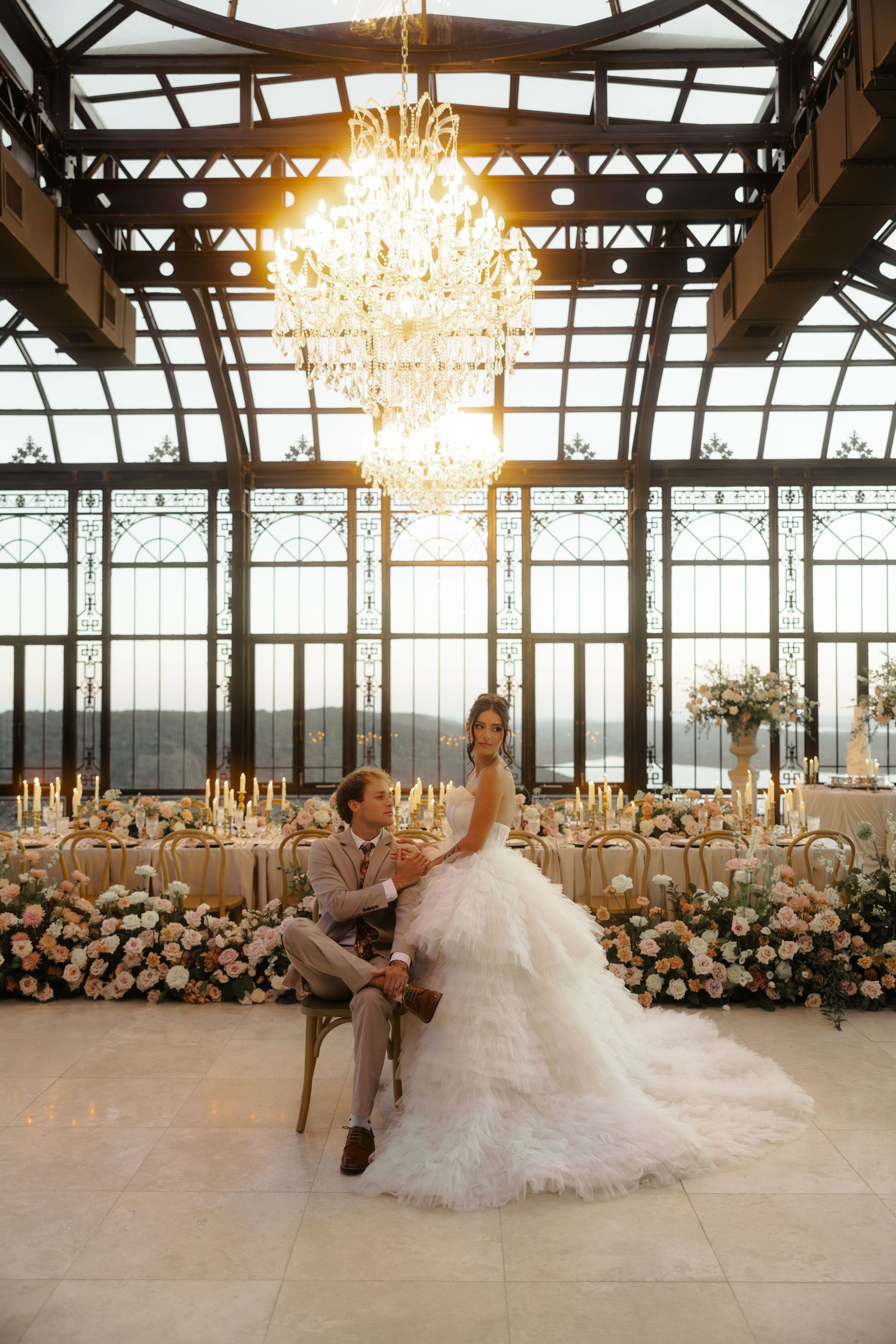 Bride and groom sit together under a grand chandelier in an elegant glass conservatory wedding venue with floral decor.