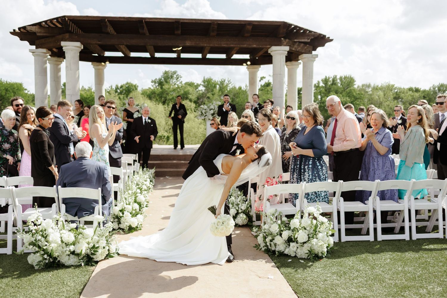 Bride and groom share a romantic dip kiss at outdoor wedding ceremony with white floral arrangements and guests applauding.