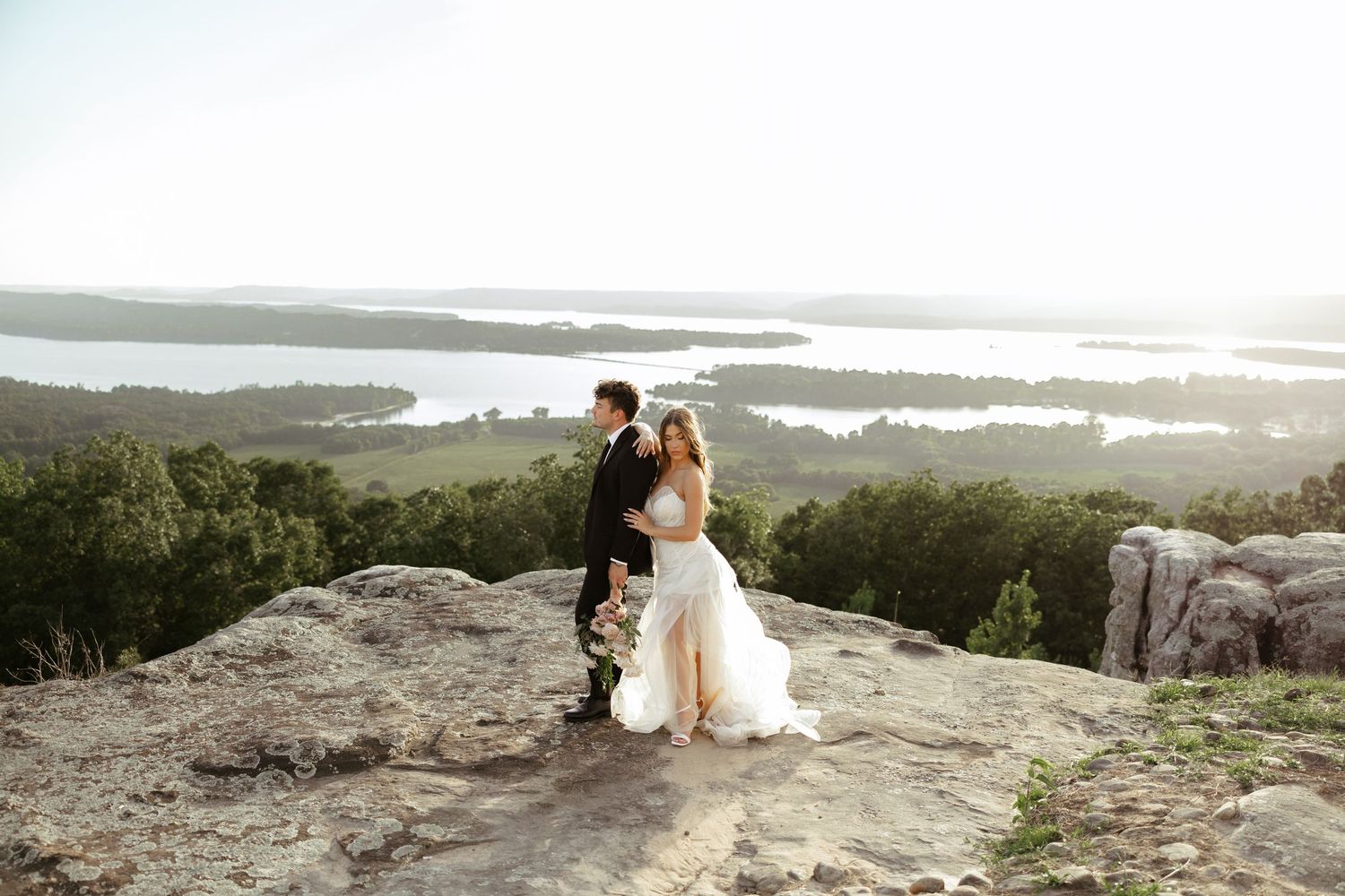 Bride and groom embrace on rocky mountaintop overlook with scenic lake and green landscape views on their wedding day.