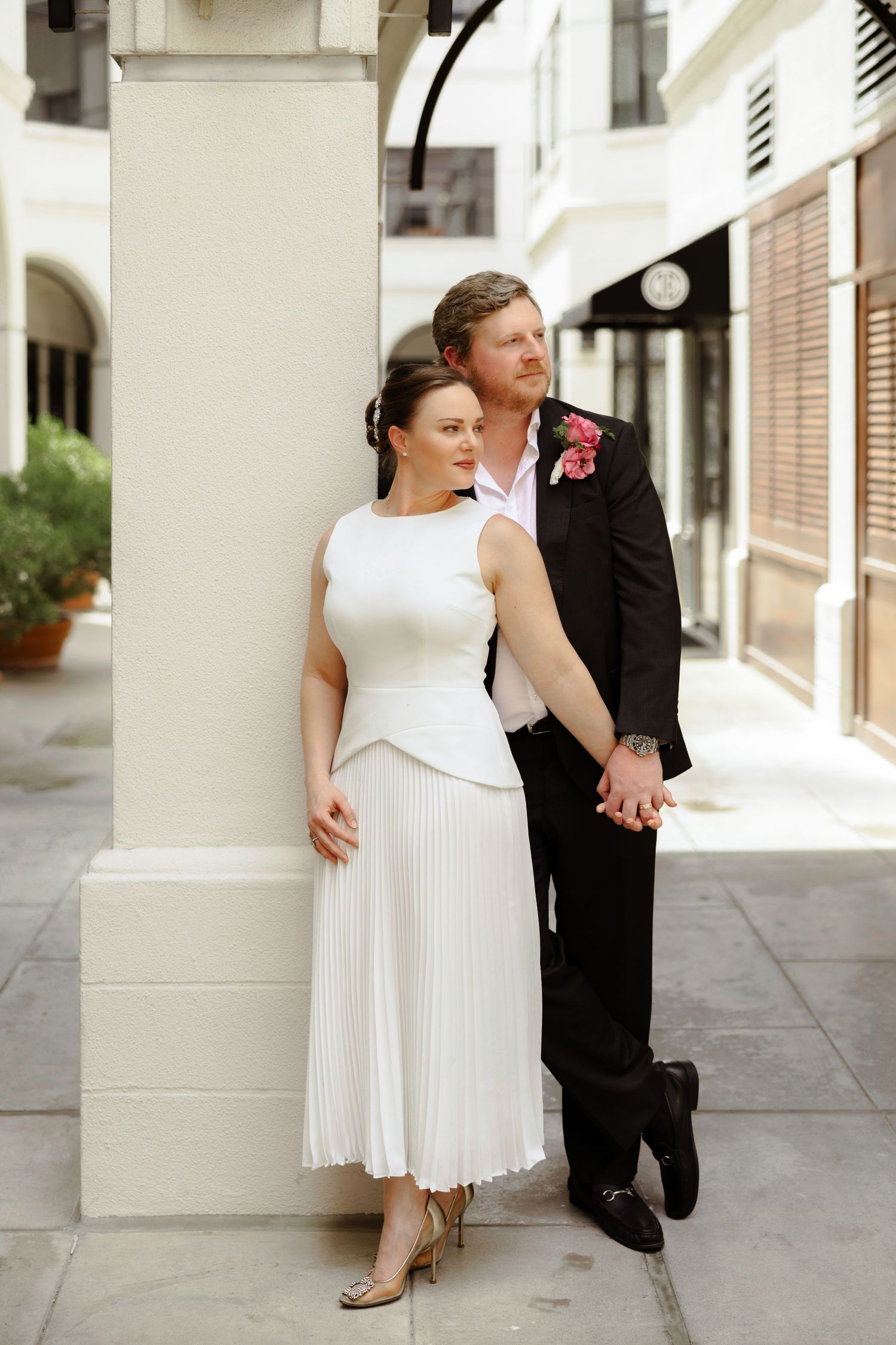 Elegant couple in wedding attire pose by a white column, bride in pleated midi dress, groom in black suit with pink boutonniere.