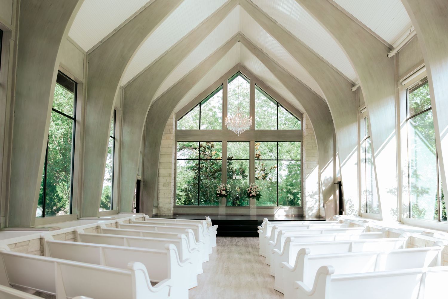 Interior of a modern white chapel with vaulted ceilings, glass windows and white pews leading to an altar.