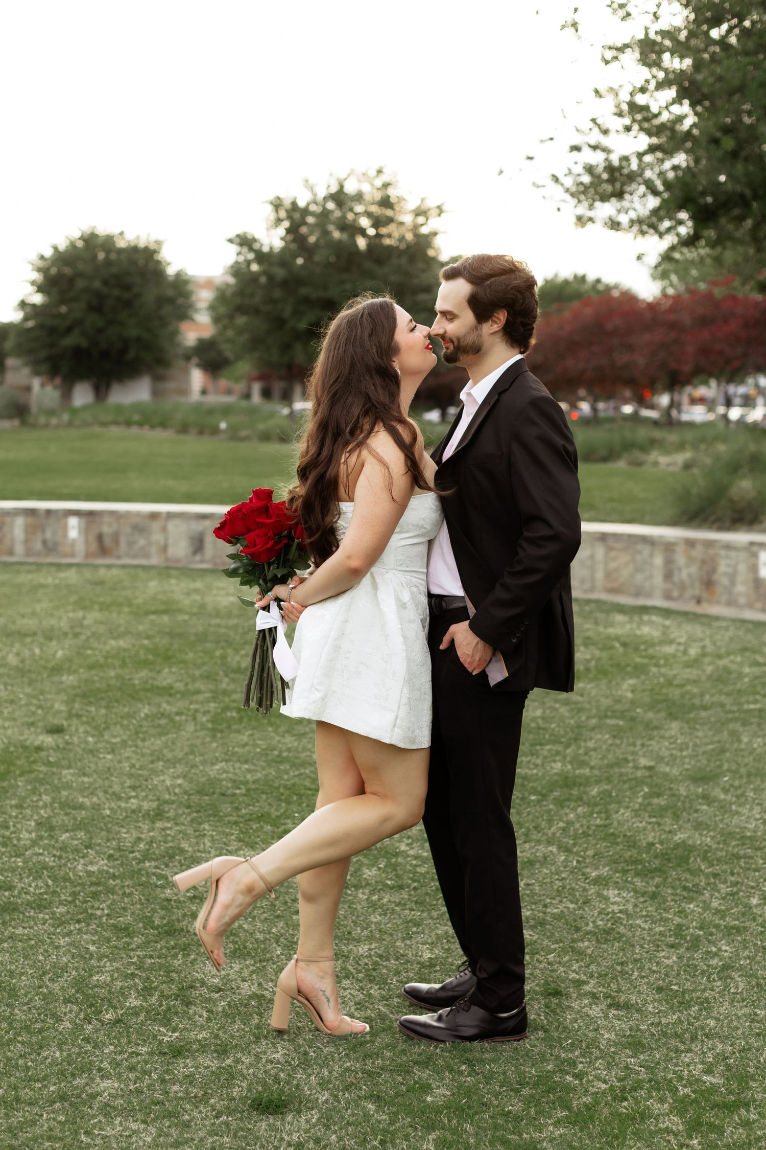 Couple kissing outdoors, woman in white mini dress holding red roses, man in black suit, standing on green grass.