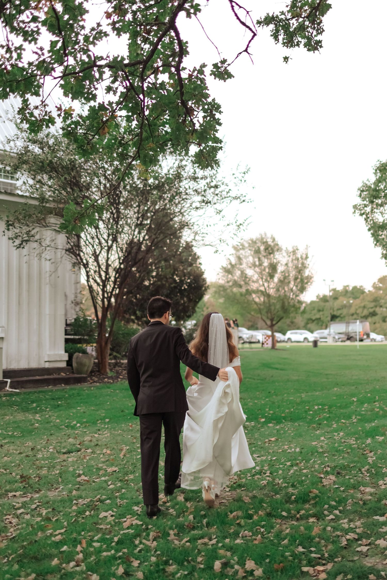 A couple walks together on a grassy lawn covered in autumn leaves near a white building.