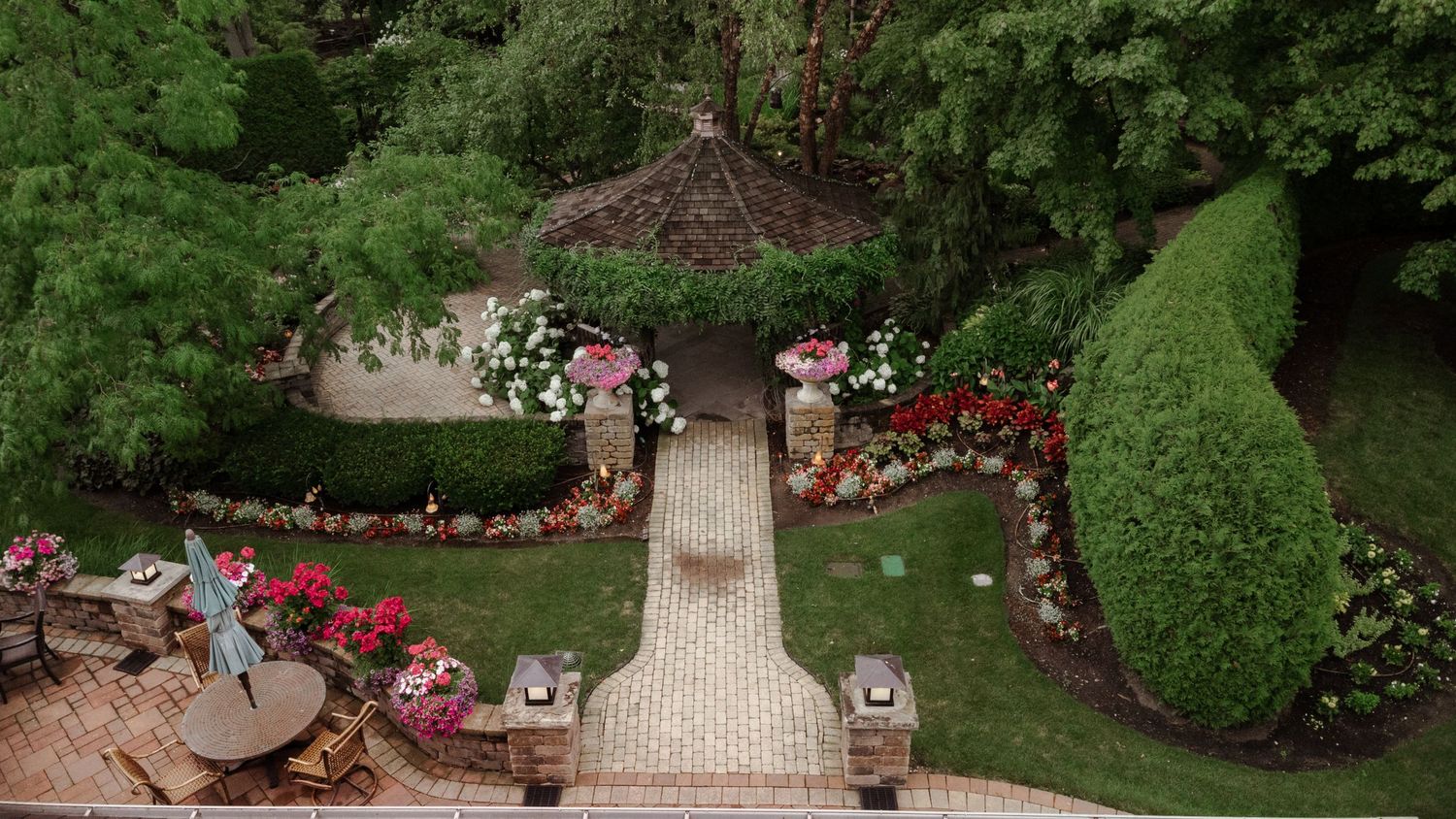 Aerial view of a lush garden with brick pathway leading to a gazebo surrounded by colorful flowers and manicured hedges.
