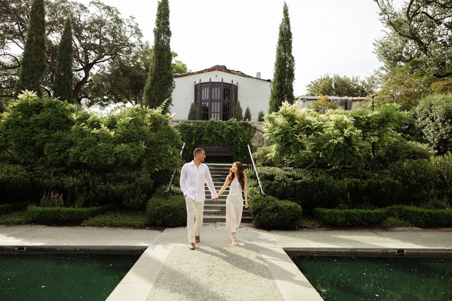 Couple in white attire walking by a pool at a lush green estate with cypress trees and Spanish villa.