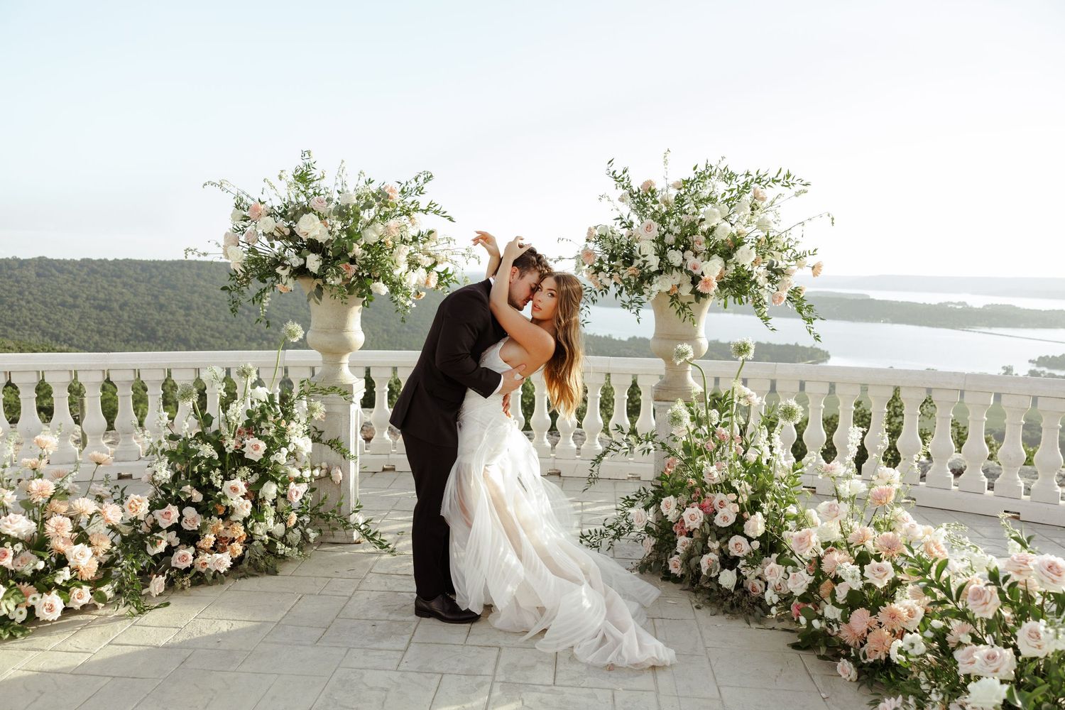 Bride and groom share a romantic kiss on a floral-decorated terrace overlooking a scenic coastal view.