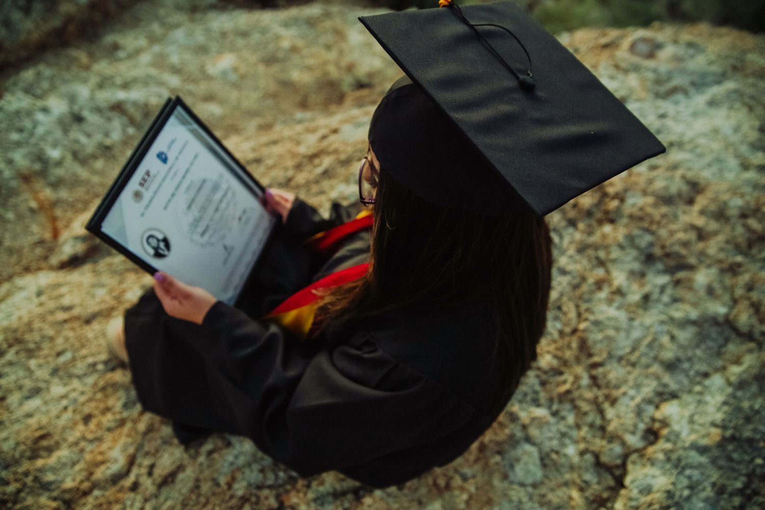 Graduate in cap and gown sits on rocks reviewing diploma certificate outdoors.