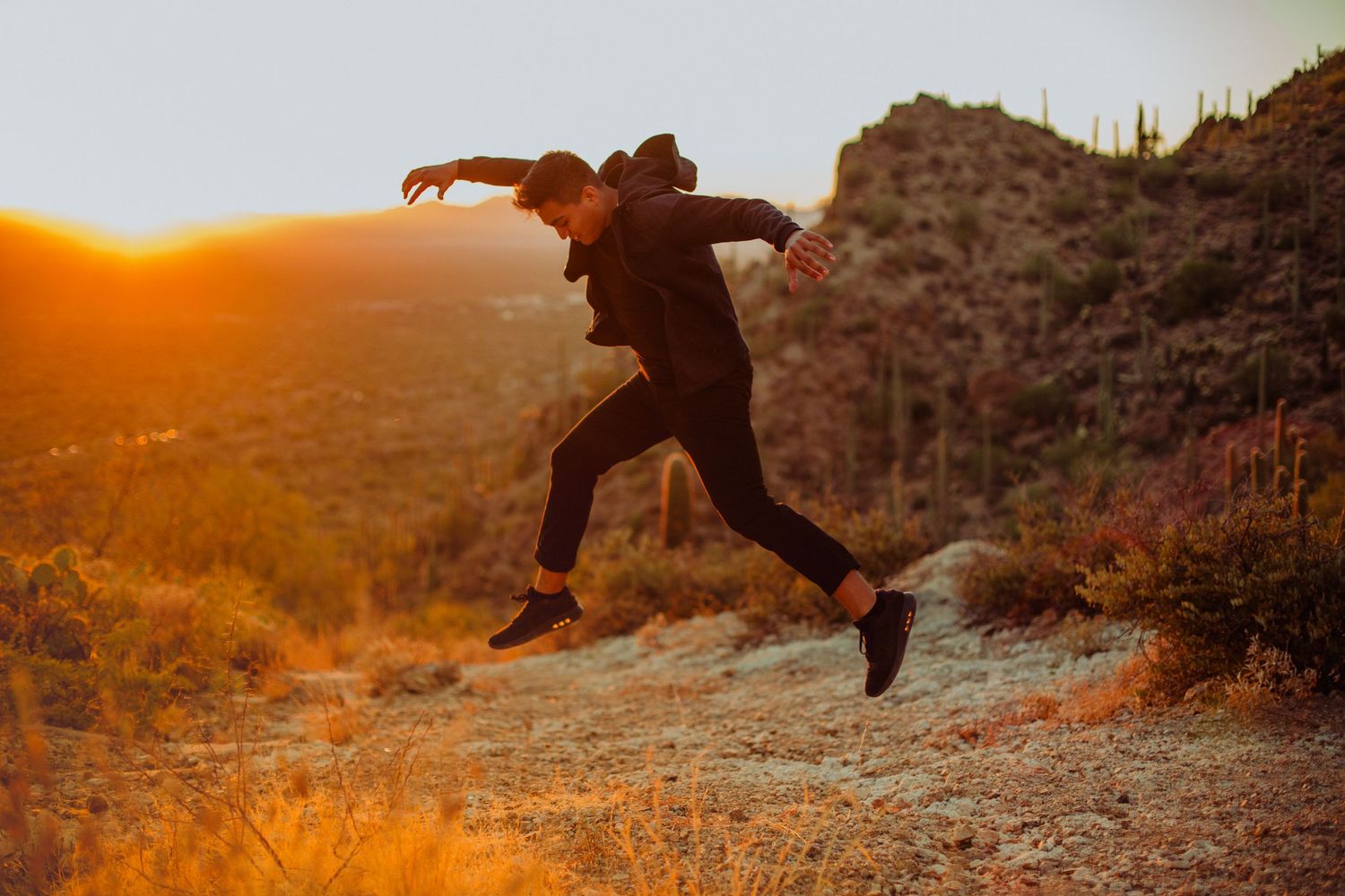Person in black outfit leaping mid-air on a rocky desert trail at golden sunset with cacti and hills in background.