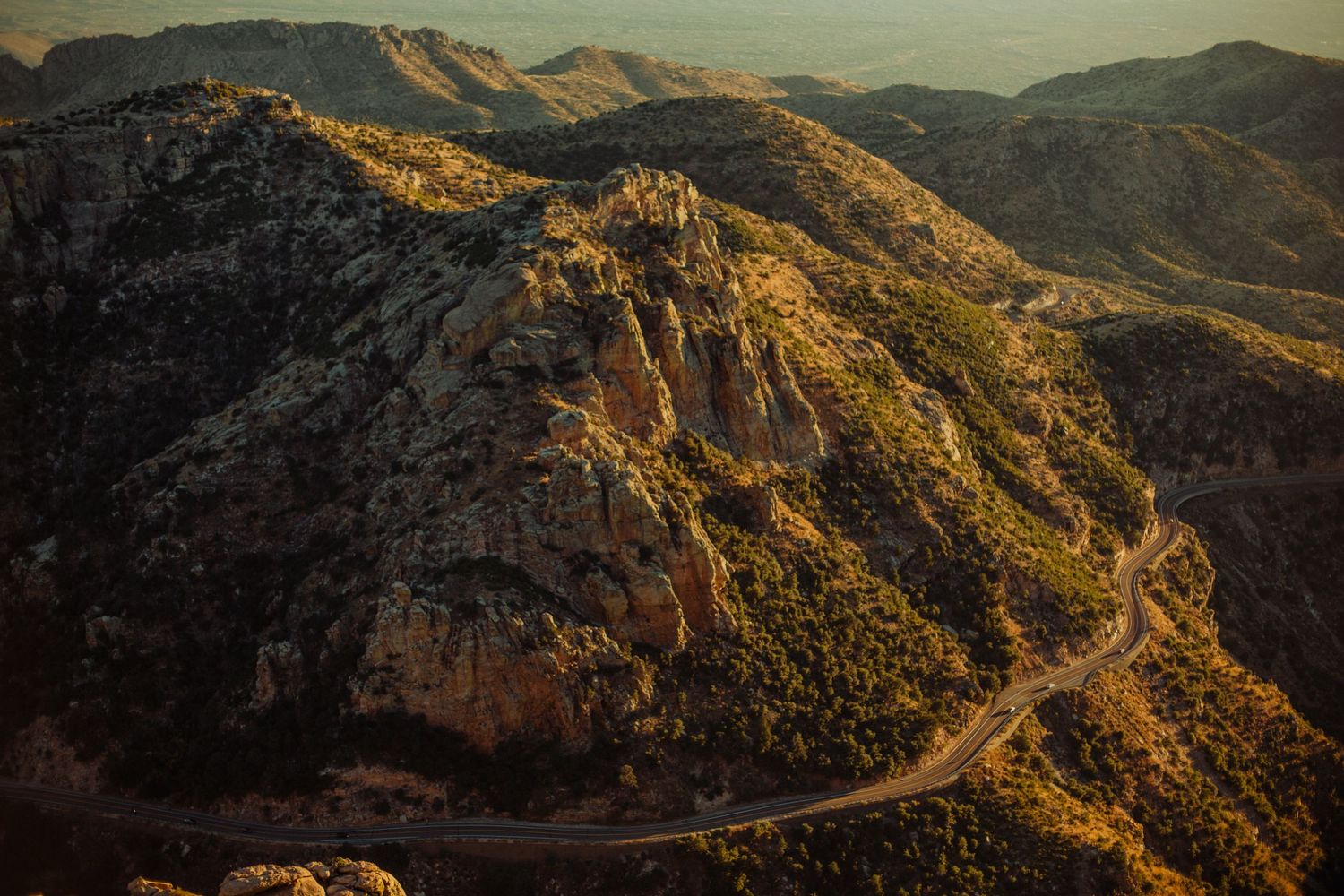 Aerial view of a winding mountain road cutting through rocky terrain bathed in warm golden sunset light.