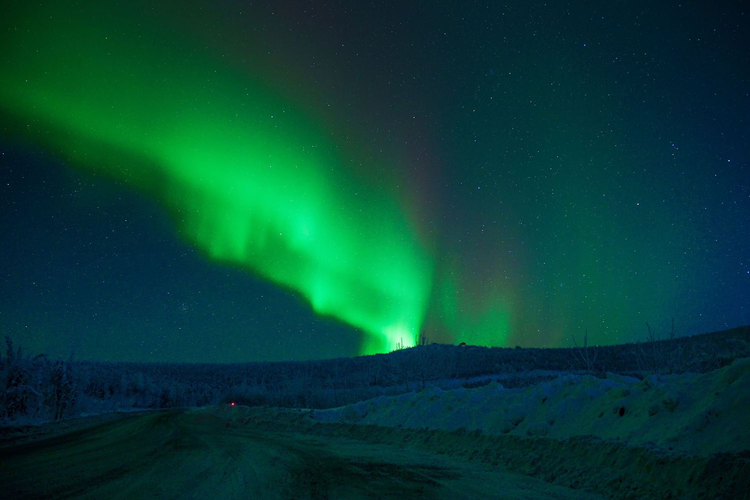 Vivid green aurora borealis illuminates a snowy winter landscape along a quiet road under a starry night sky.