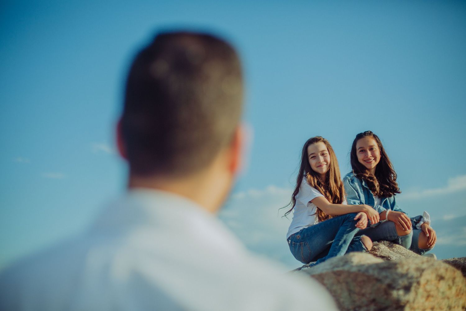 Two teenage girls sitting on rocks smiling at a man in a white shirt against a blue sky backdrop.