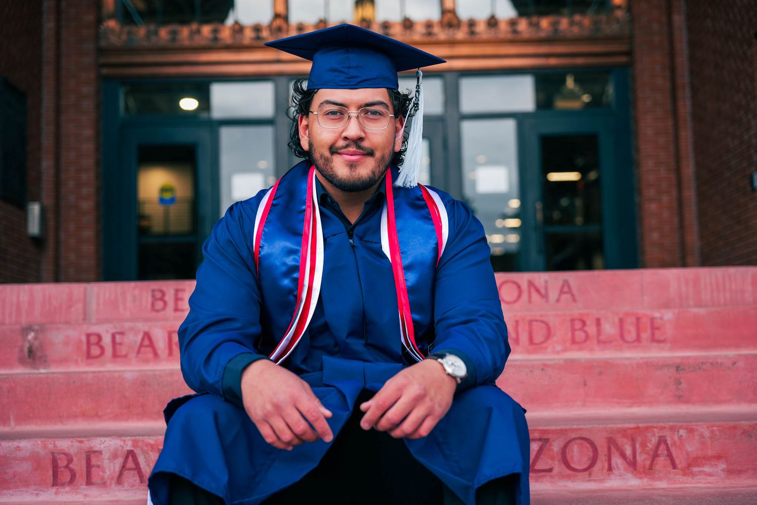 College graduate in blue cap and gown sits on University of Arizona steps, smiling proudly on graduation day.