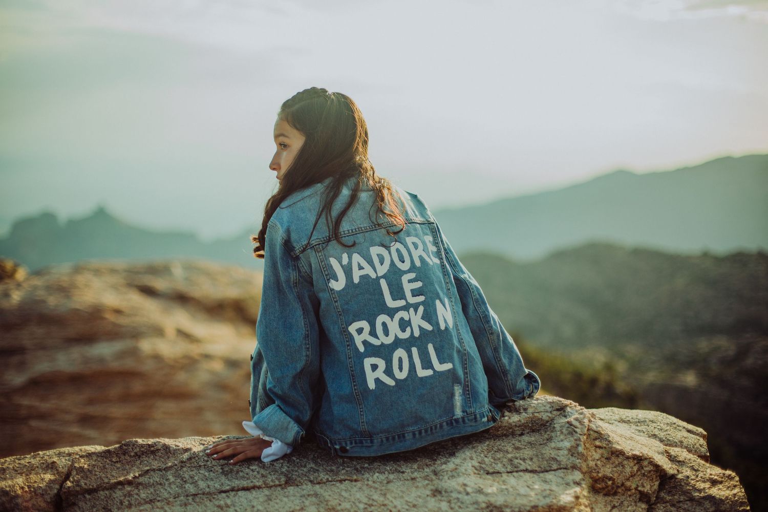 Woman in denim jacket with J'adore Le Rock N Roll text sits on rocky mountain overlook at sunset.