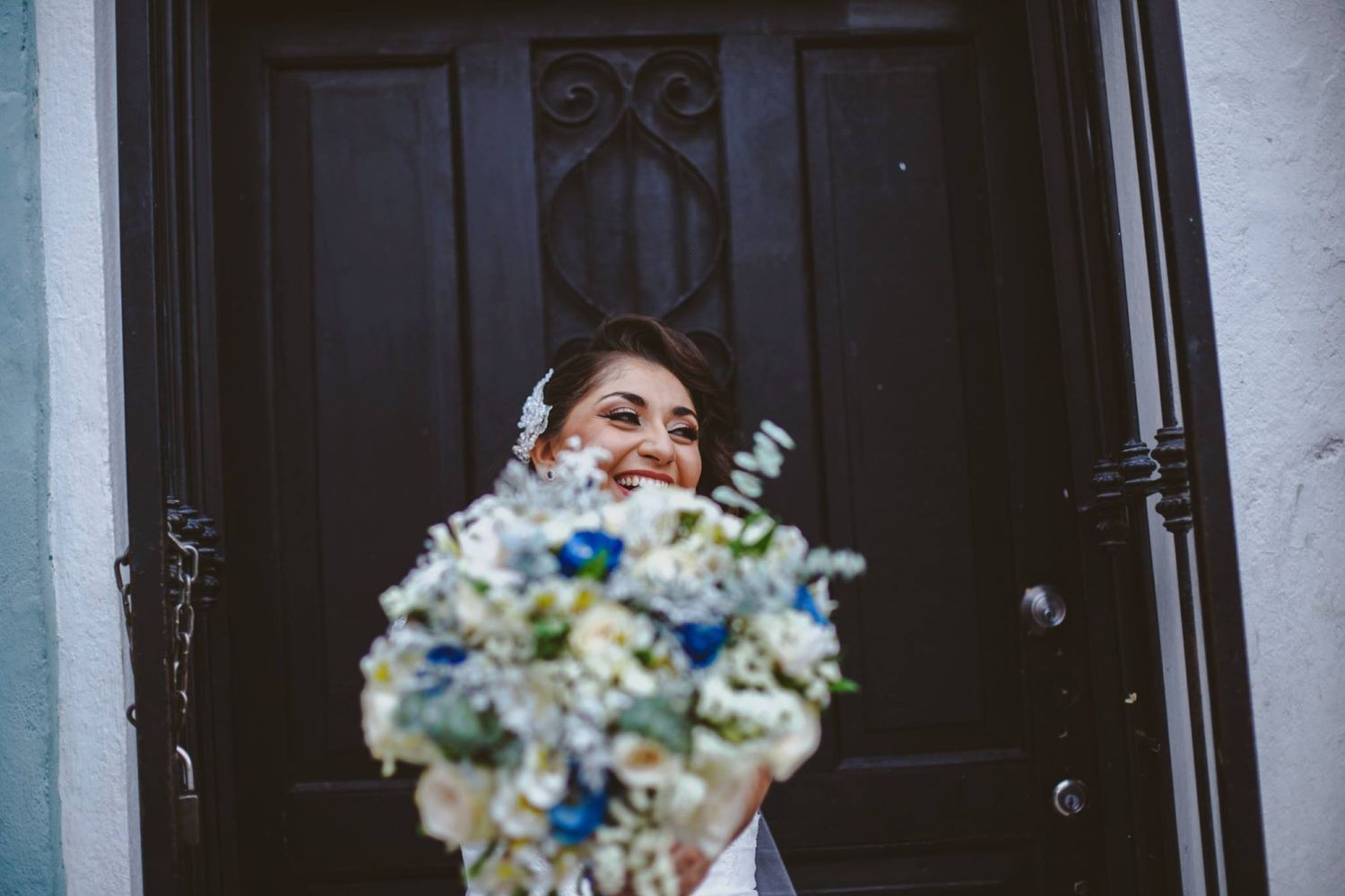 Smiling bride holding a white and blue floral bouquet in front of a dark wooden door on her wedding day.